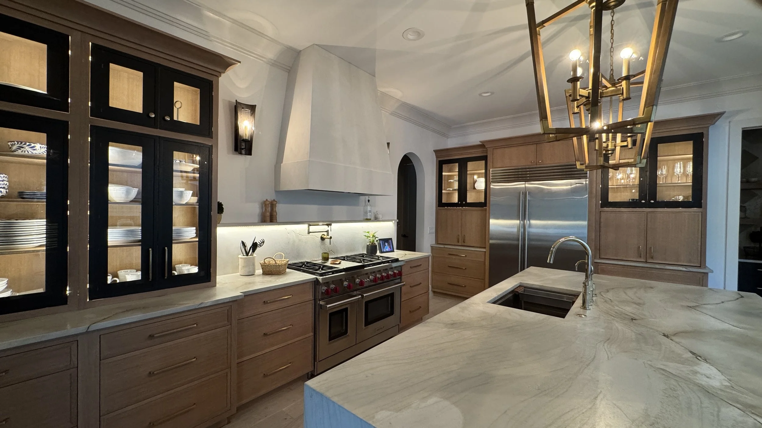 Modern kitchen with light wood cabinets, marble countertops, stainless steel appliances, and a gold chandelier