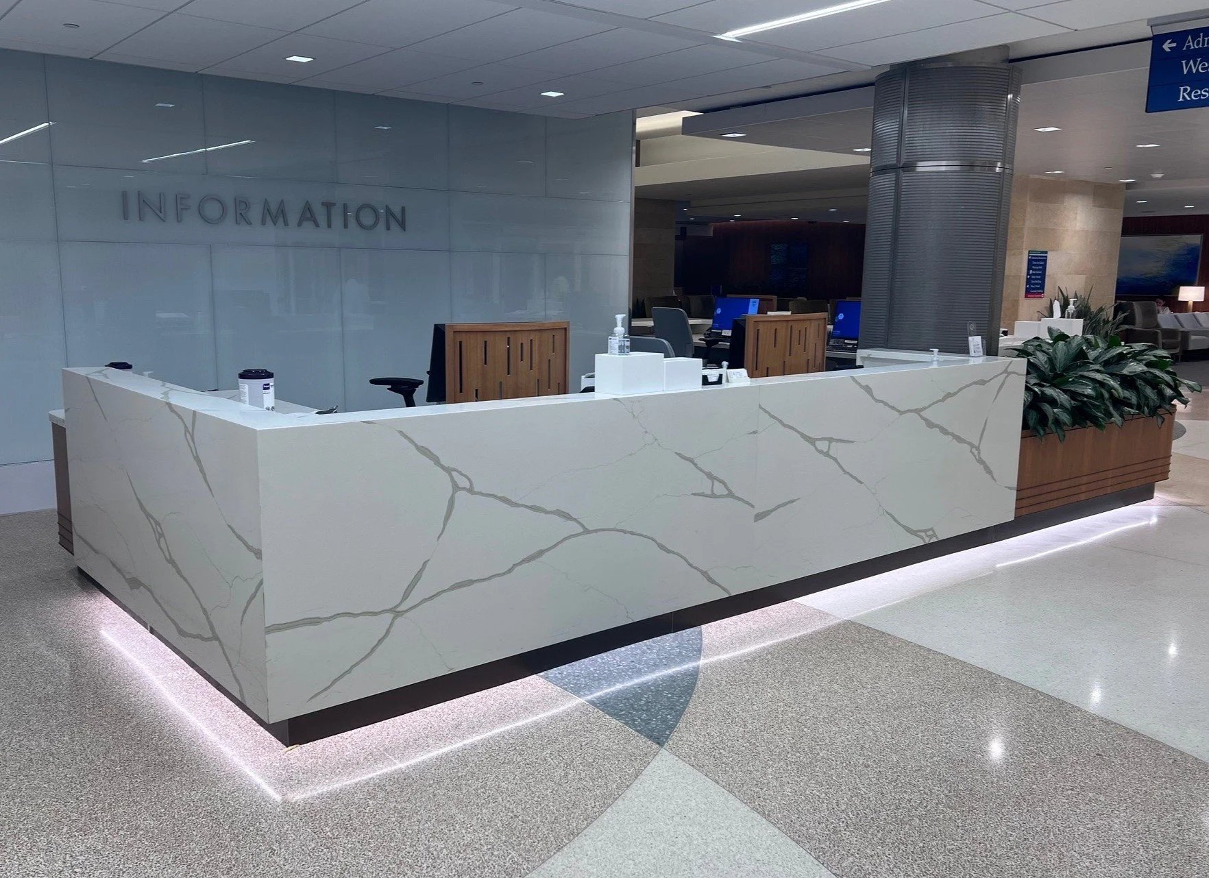 An airport information counter with a white marble counter, computer monitors, and hand sanitizer, located in a modern terminal with plants and seating areas in the background.