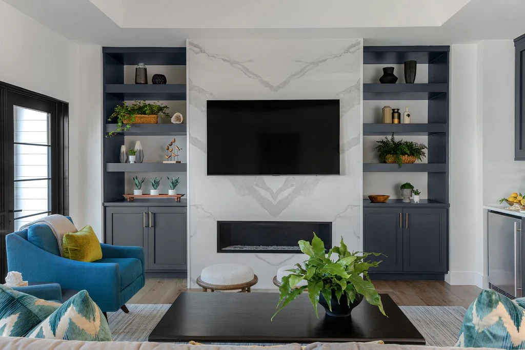 Living room with a wall-mounted TV above a fireplace, blue built-in shelves on each side with decorative items, a black coffee table with a green plant, blue armchair with a yellow pillow, and windows with black trim.