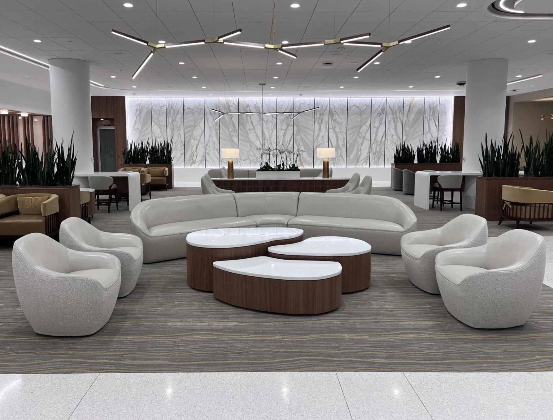 Modern hotel lobby with white curved sofas and armchairs arranged around wooden and white tables, a marble wall with backlighting, and stylish ceiling lights.