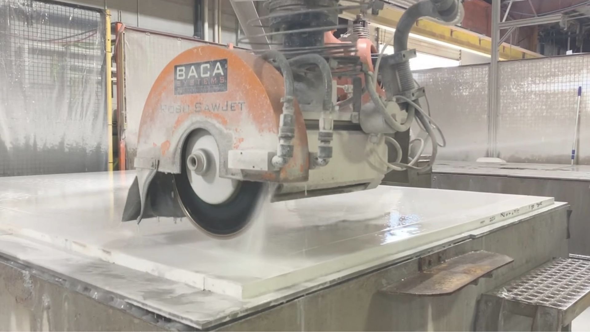 A large industrial circular saw cutting through a thick slab of material in a factory setting with protective fencing and machinery in the background.