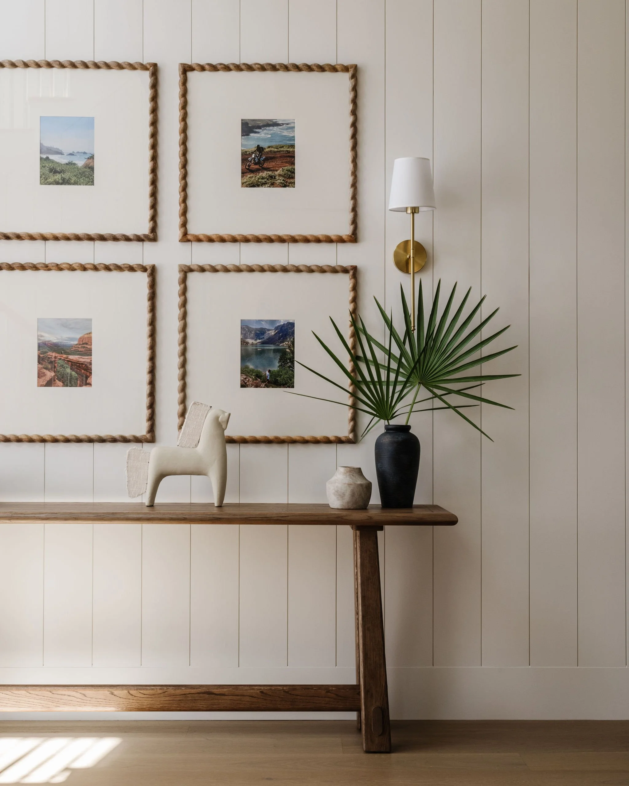 Interior of a room with wooden flooring and a wall decorated with framed landscape photos, with thick rope borders around the frames. A white chair with beige upholstery partially visible in front, a wooden table, a tall black vase with a large green