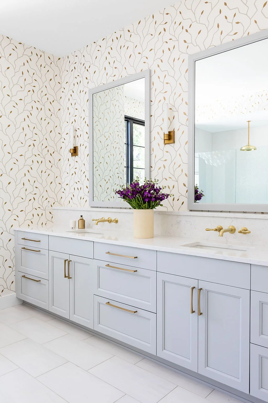 Modern bathroom with white vanity, gold handles, gold fixtures, large mirrors, floral wallpaper, window, and purple flower arrangement.