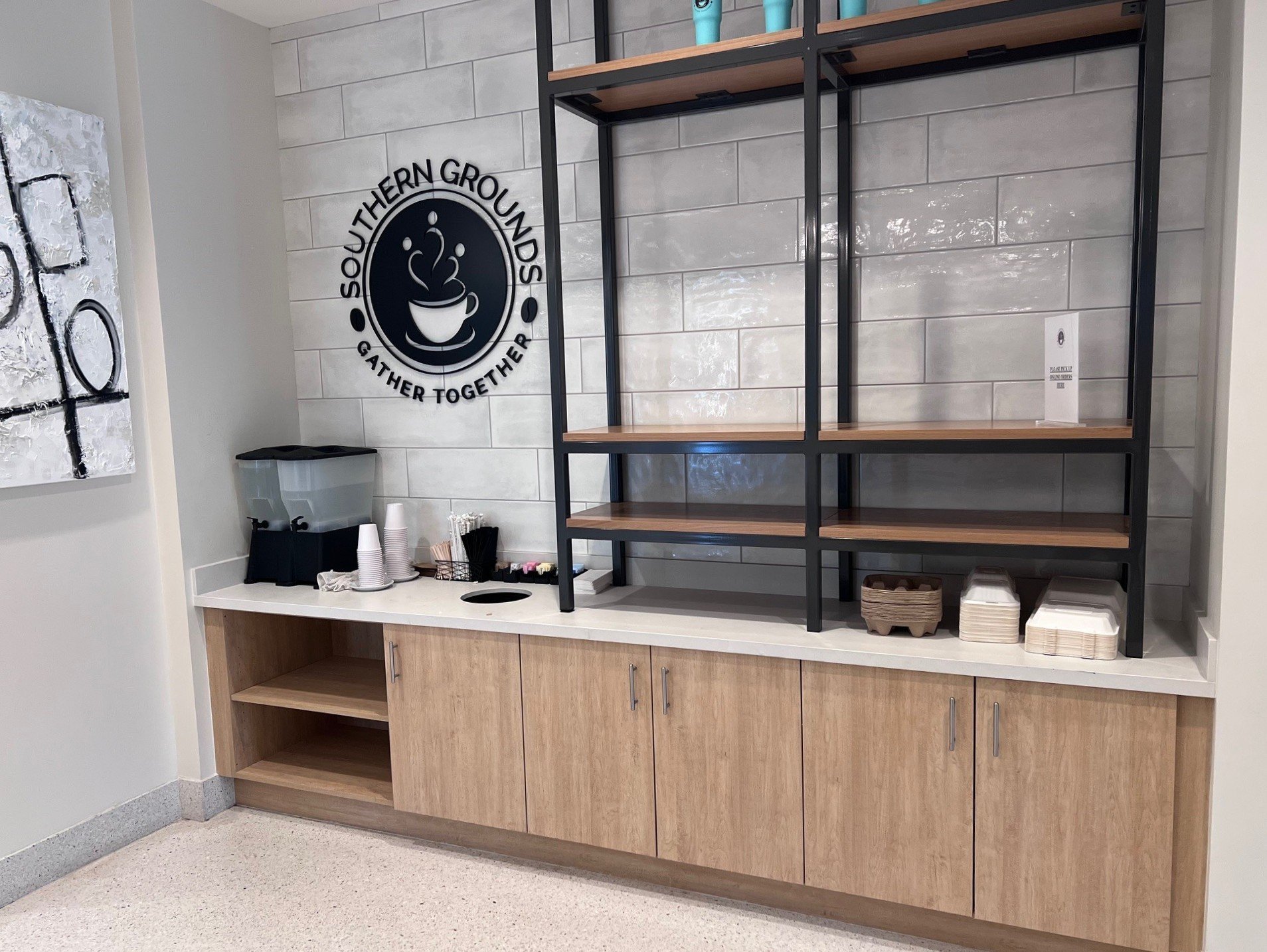 Empty coffee station with a black metal shelf in a modern cafe or office setting. The shelf has three wooden shelves on a tiled wall with light-colored tiles. Below the shelf is a counter with disposable cups, stir sticks, and a napkin dispenser. To 