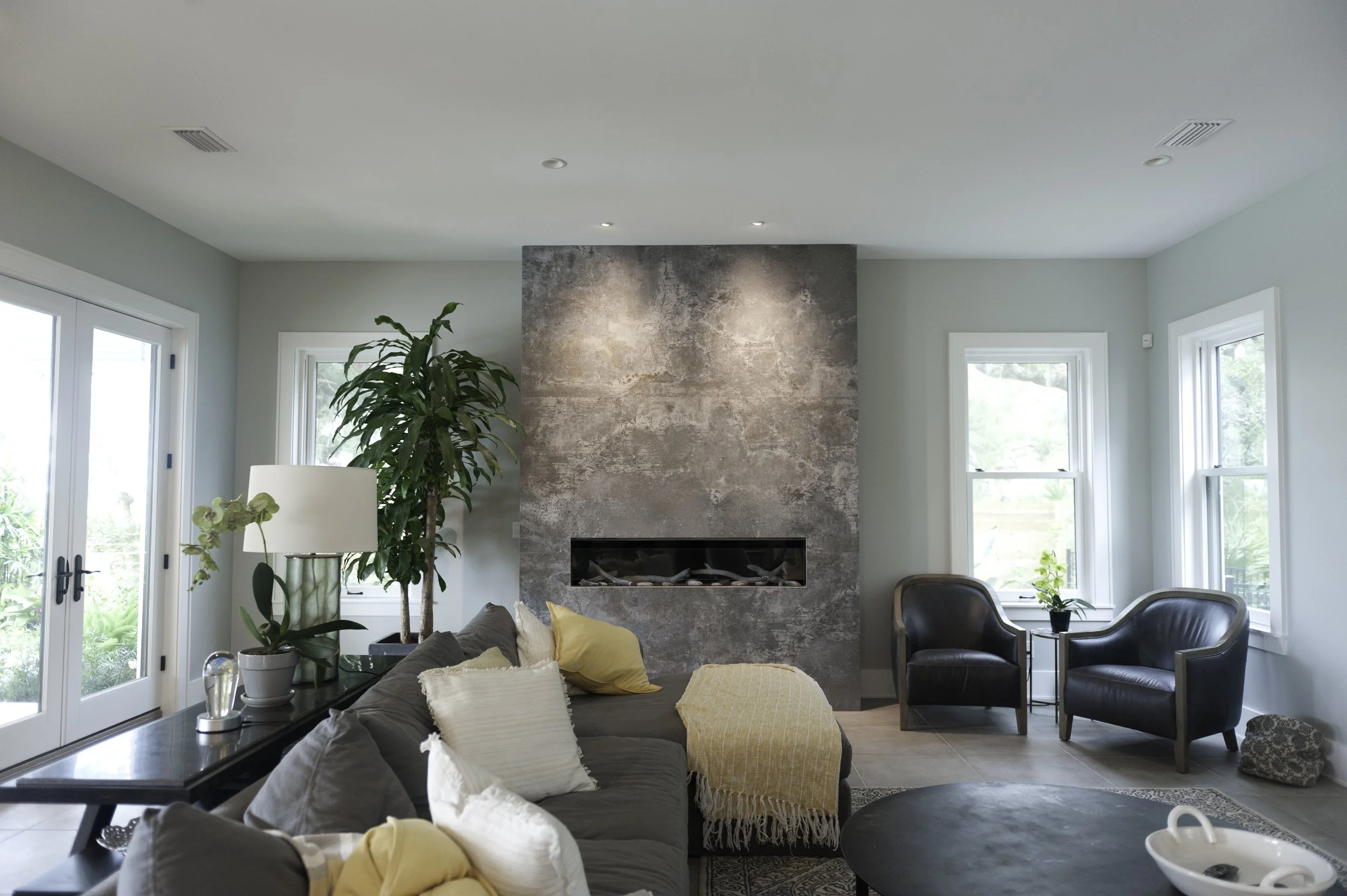 Living room with a stone fireplace, large windows, green plants, a gray sofa with throw pillows, a black armchair, and sunlight streaming in.