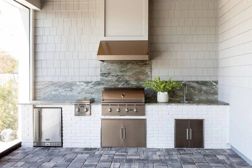Outdoor modern kitchen with stainless steel grill, sink, cabinets, a potted plant, and a stone and brick backsplash, located next to a glass sliding door.