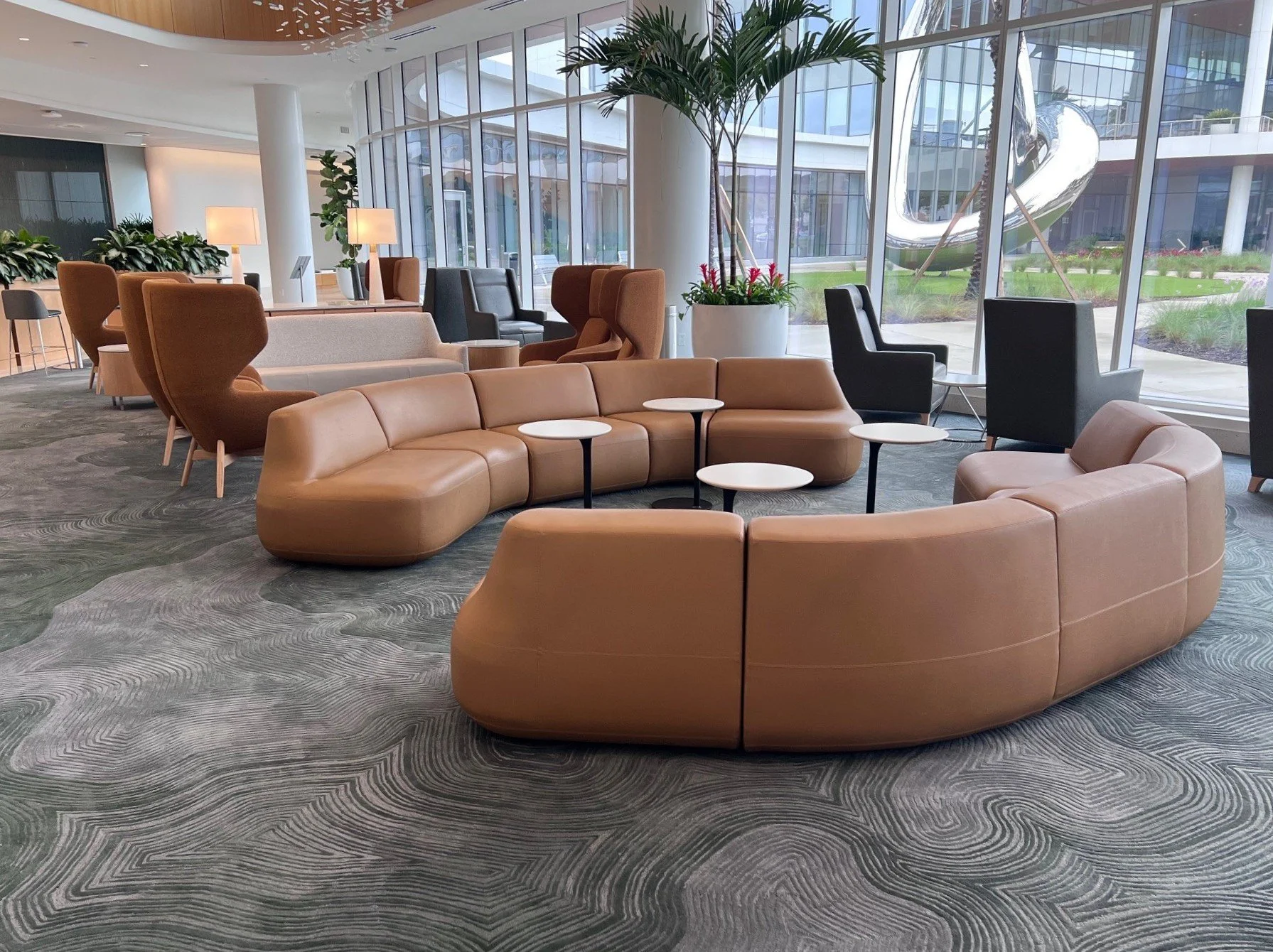 Modern airport lounge with curved tan leather sectional sofa, black and brown accent chairs, small side tables, large potted plants, and floor-to-ceiling windows showing a sculpture outside.