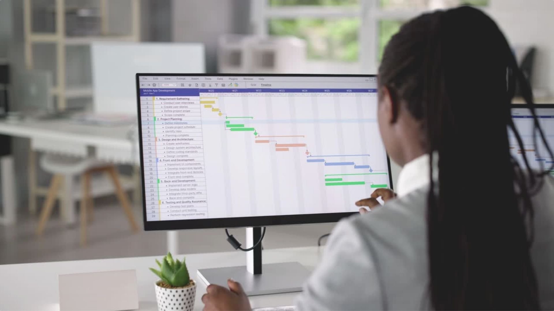 A person working on a computer with a large monitor displaying a colorful project management Gantt chart in a bright, modern office.