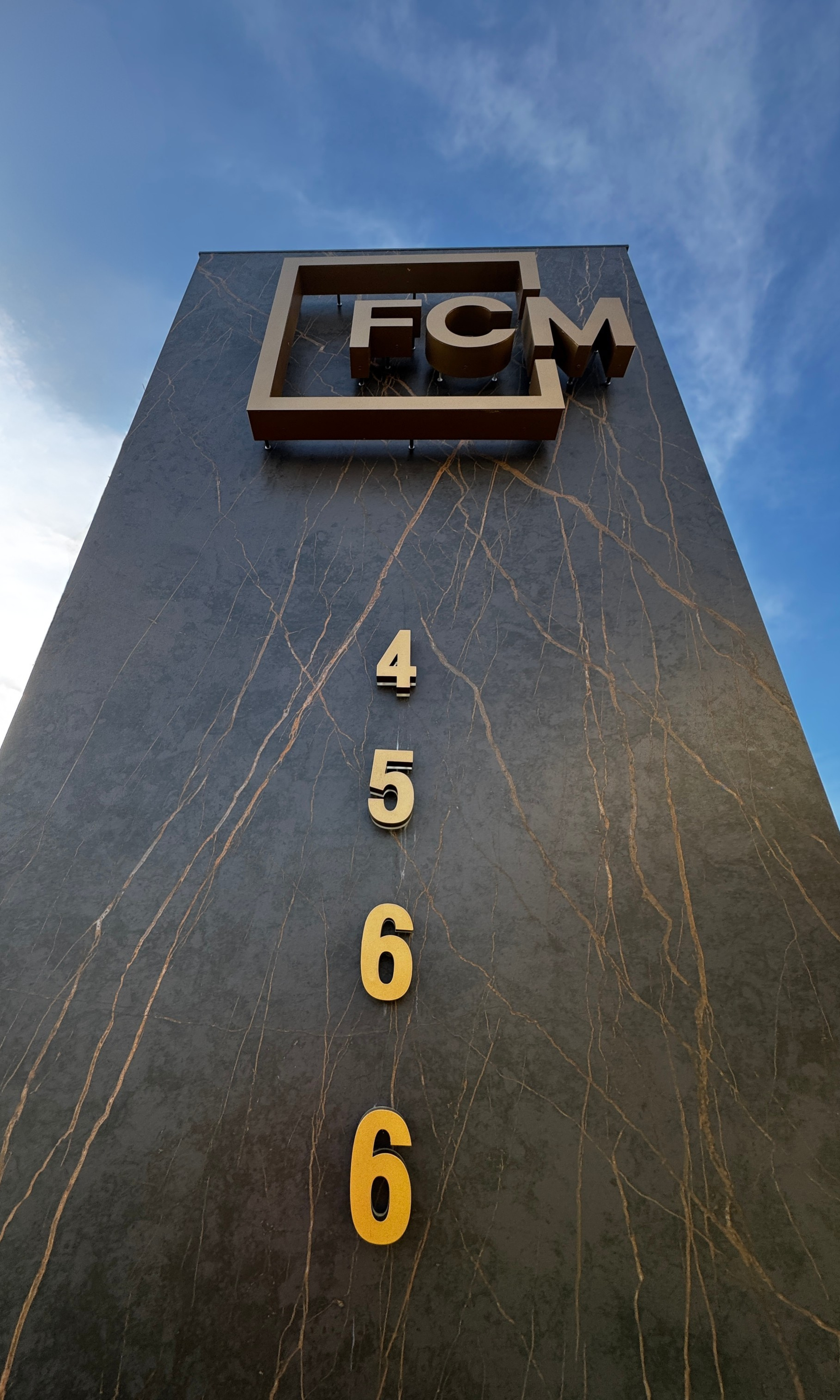 A tall, dark marble monument with a square logo bearing the letters 'FCM' at the top and large gold-colored numbers from 4 to 6 running vertically down its front. The sky is blue with some clouds.
