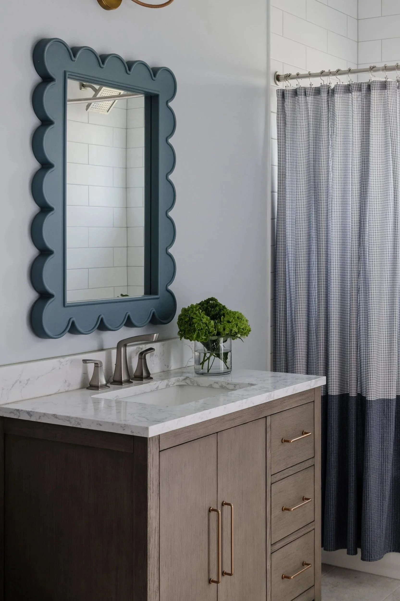 A bathroom vanity with a white marble countertop and dark wood cabinets with gold hardware. A glass vase with green hydrangeas sits on the counter. A large, decorative blue mirror hangs above the sink. To the right, there is a shower with a checkered curtain and subway tile walls.