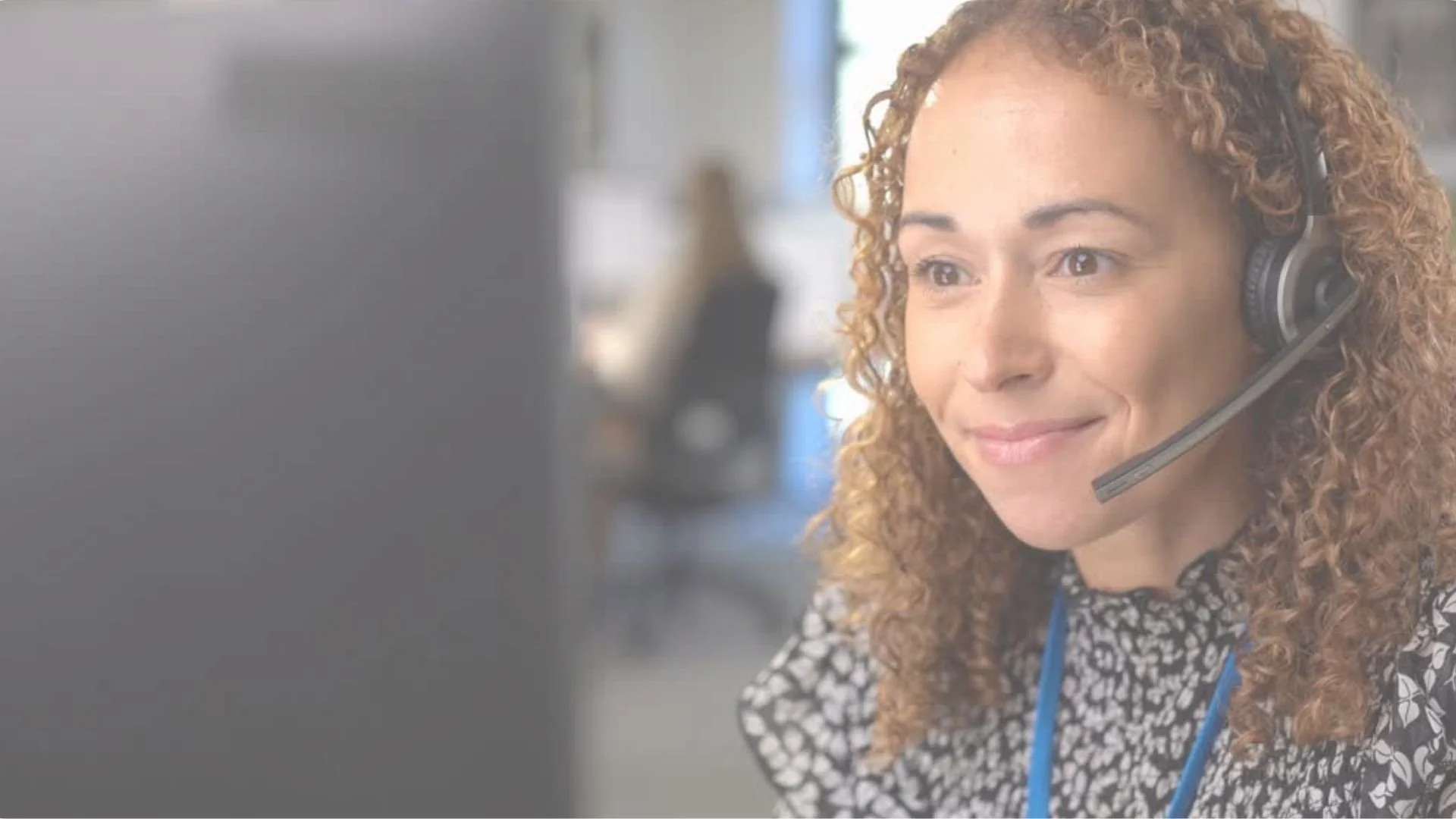 A woman with curly hair wearing a headset with a microphone, smiling while working at her desk in an office.