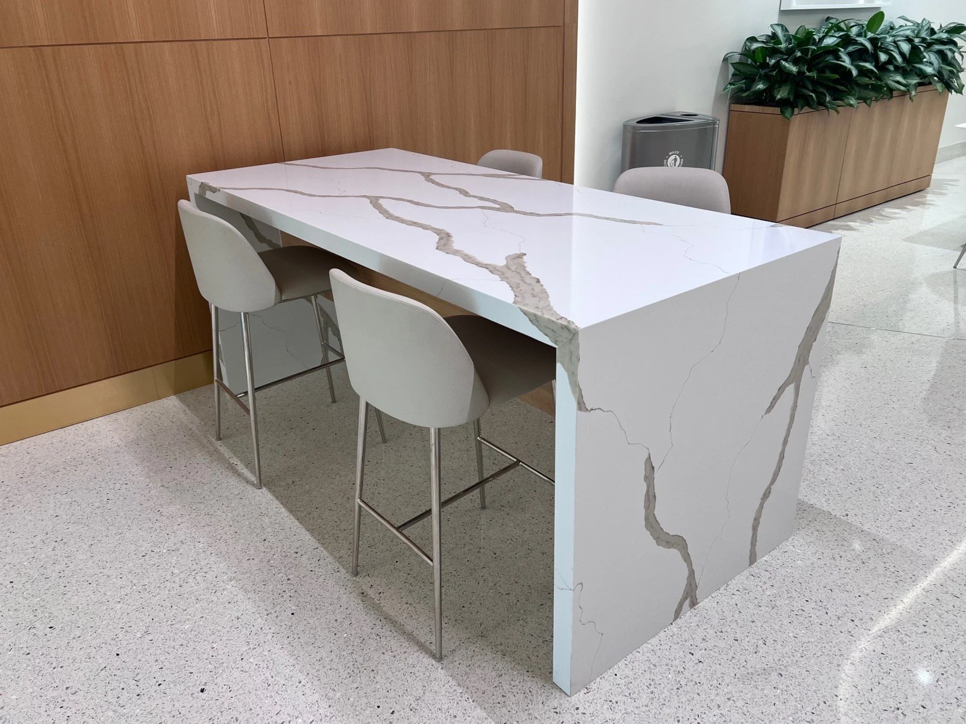 White marble table with dark veining, surrounded by light gray chairs, in an indoor area with a wooden wall, a large potted plant, and a trash bin.