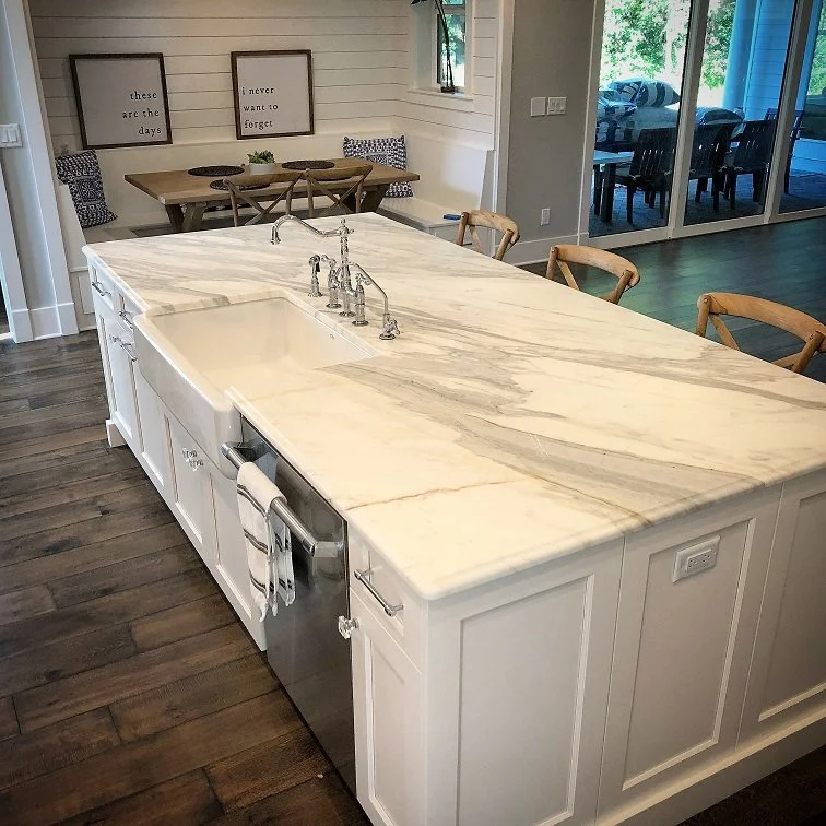 A white kitchen island with a marble top, sink, and chrome faucet, surrounded by wooden chairs, with a dining area and sliding glass doors opening outside.