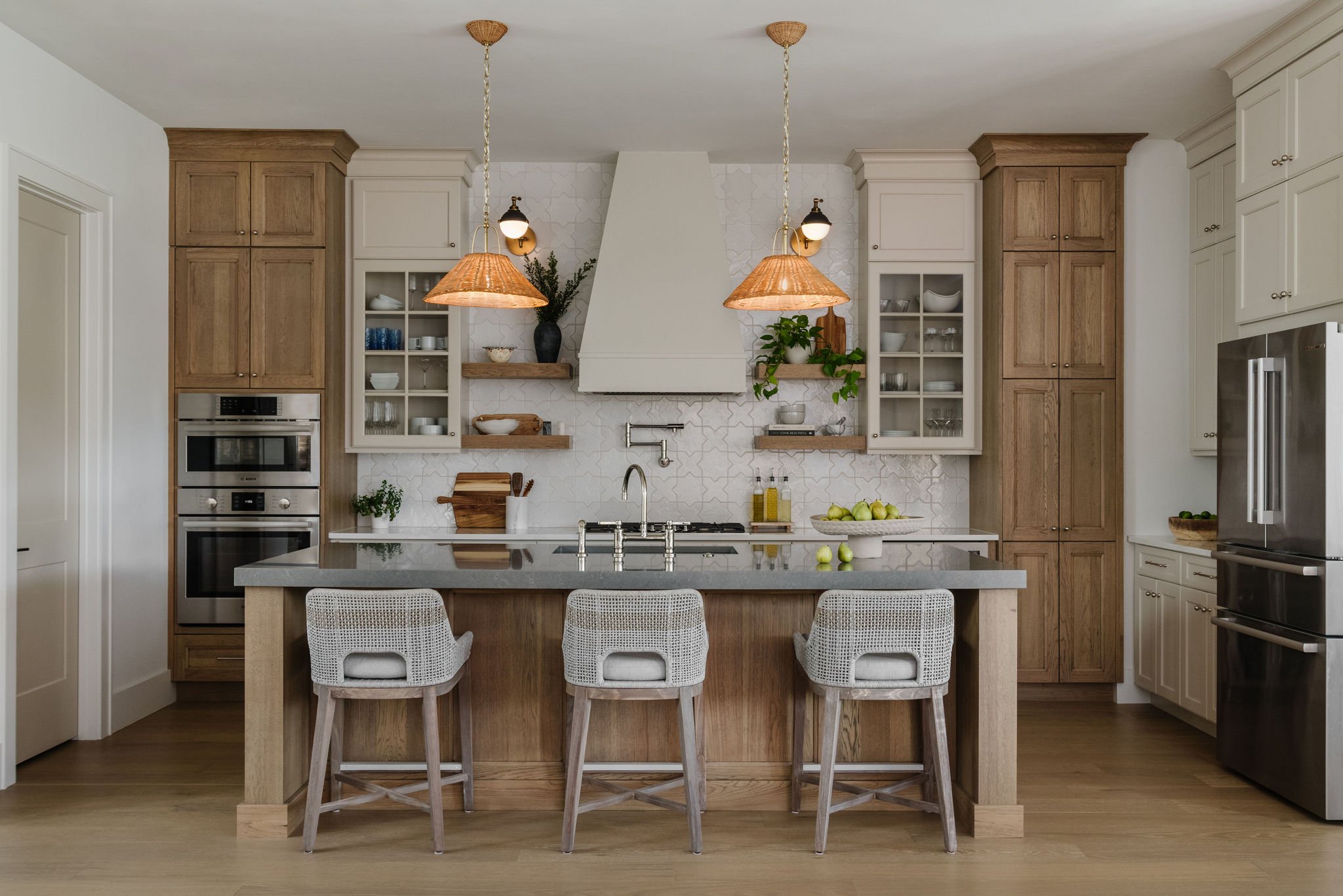 Modern kitchen with white and wood cabinets, two pendant lights, open glass cabinets with dishware, a white range hood, and a kitchen island with four white woven chairs. Green plants and apples are on the shelves.