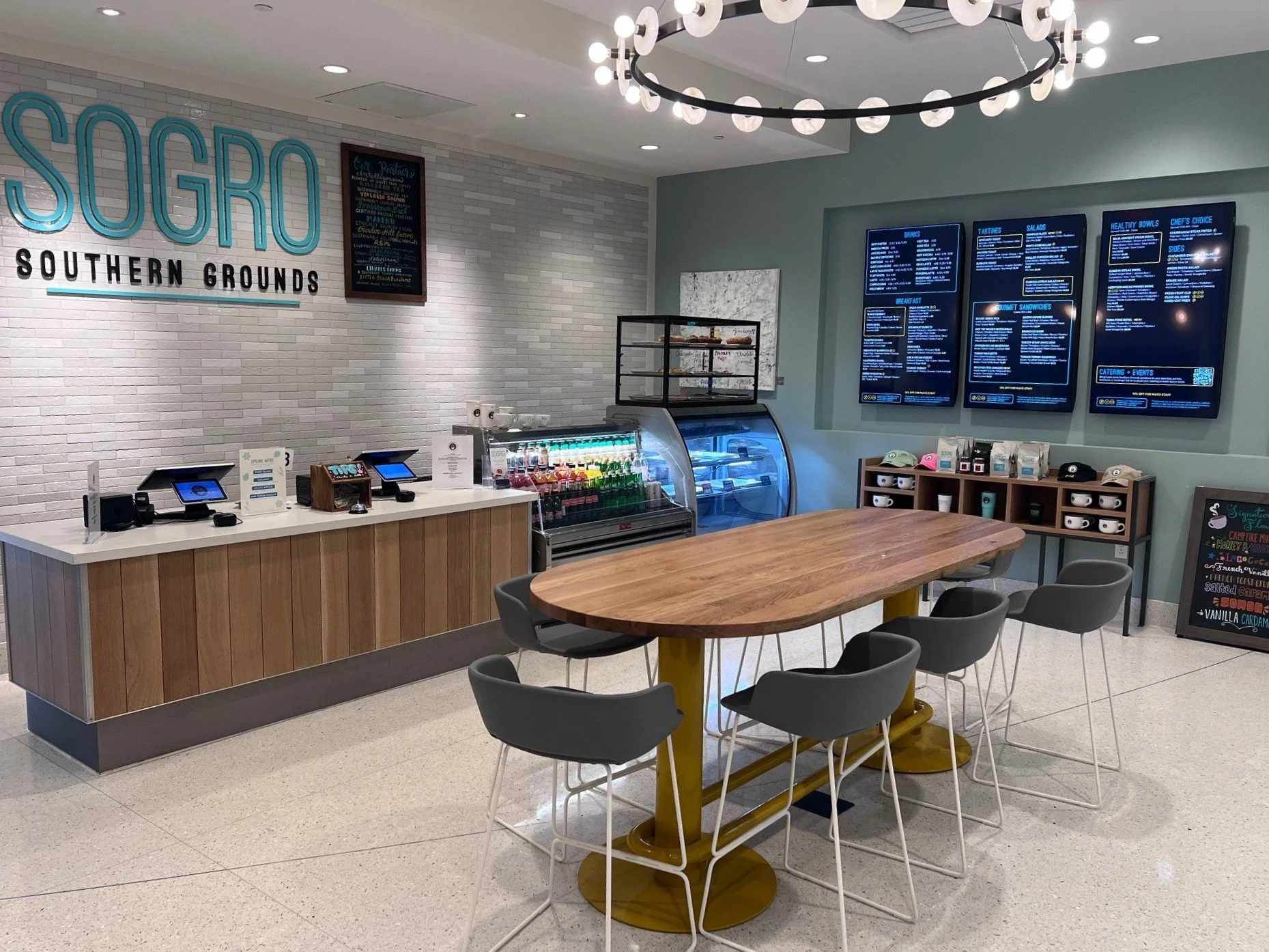 Interior of a modern restaurant or cafe with a wooden table, gray chairs, a white brick wall with a large sign that reads "SUGRO SOUTHERN GROUNDS," a blackboard menu, a digital menu board on the wall, and a refrigerated display case with drinks. Ther