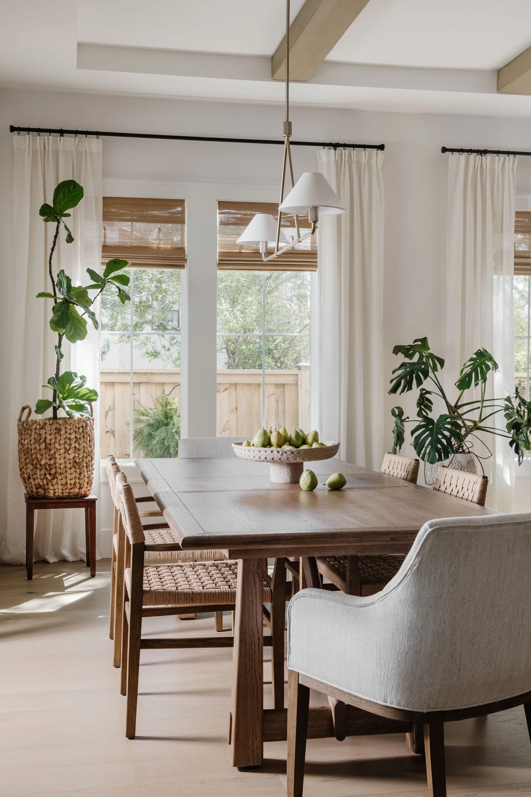 A dining room with a wooden table, woven chairs, a white armchair, potted plants, and a vase with pears, with large windows and white curtains.