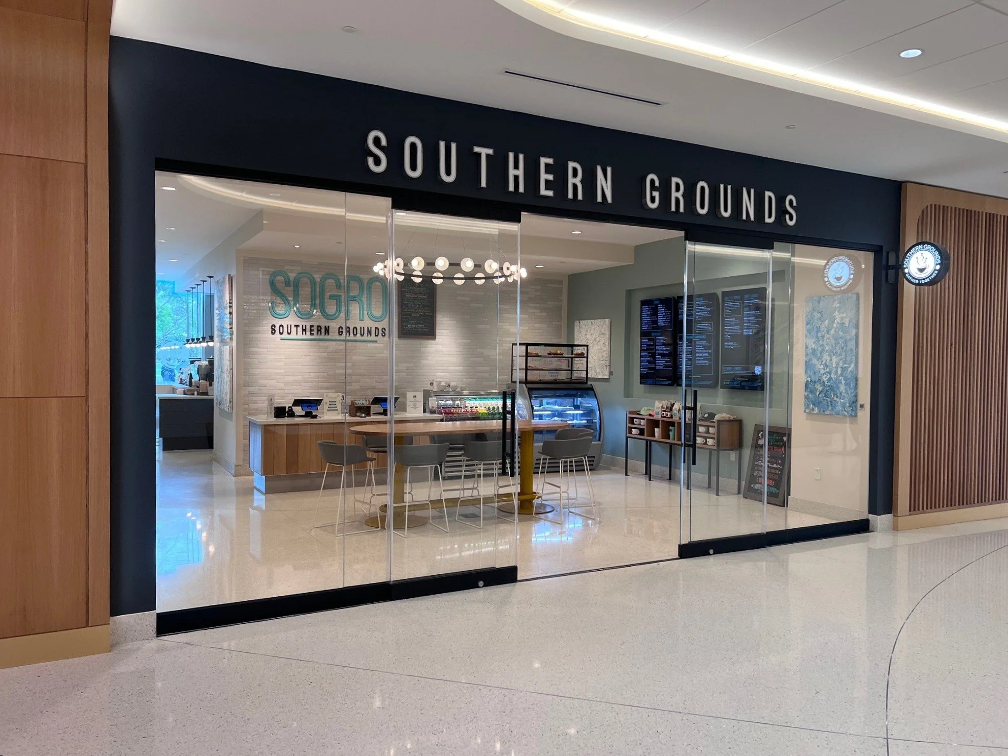 Entrance to a restaurant called Southern Grounds, with glass doors, a table with chairs inside, digital menu screens, and decorative lighting fixtures inside.