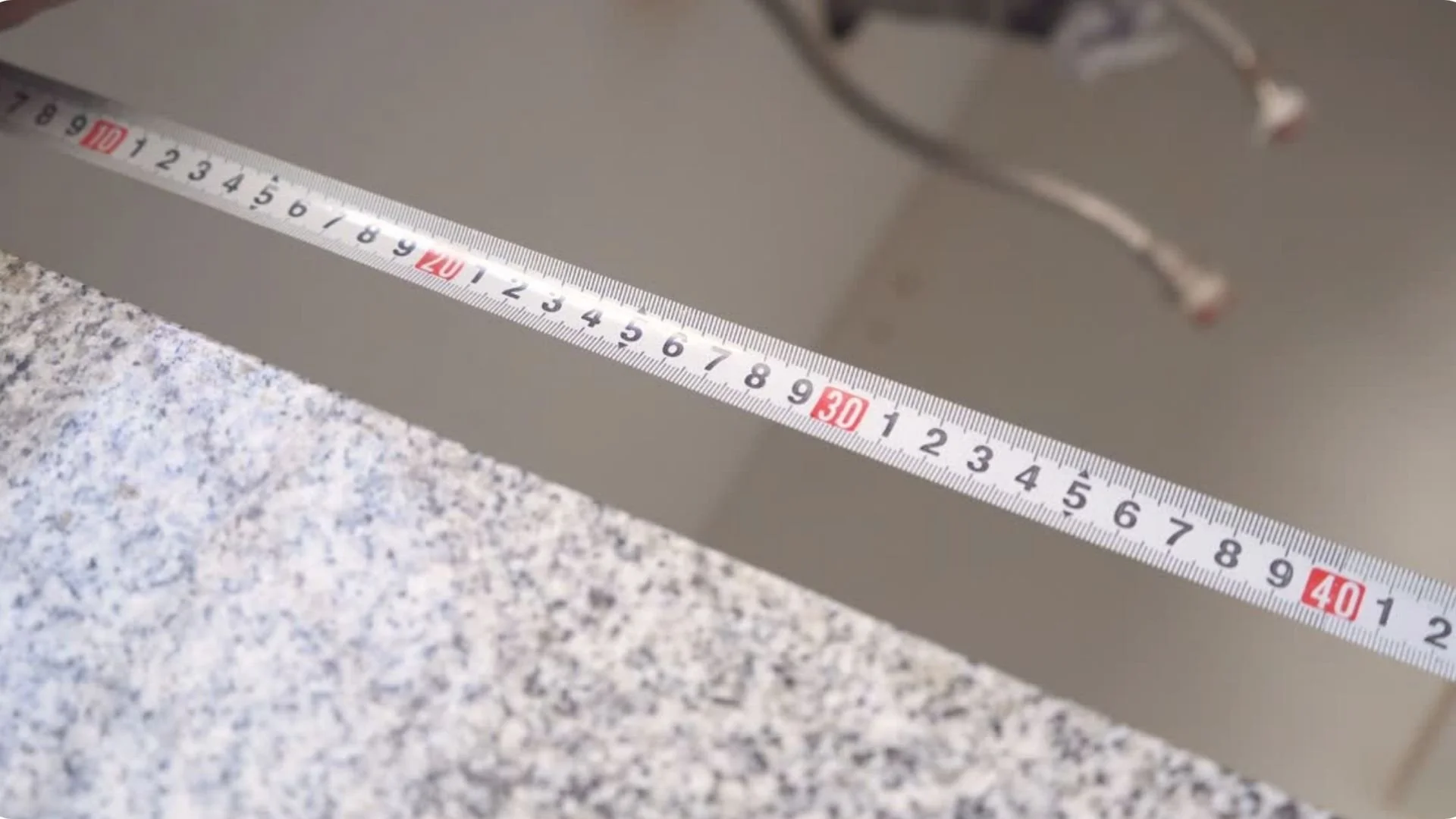 A measuring tape laid across a flat surface, with part of a metal clothing rack or hook visible in the background.