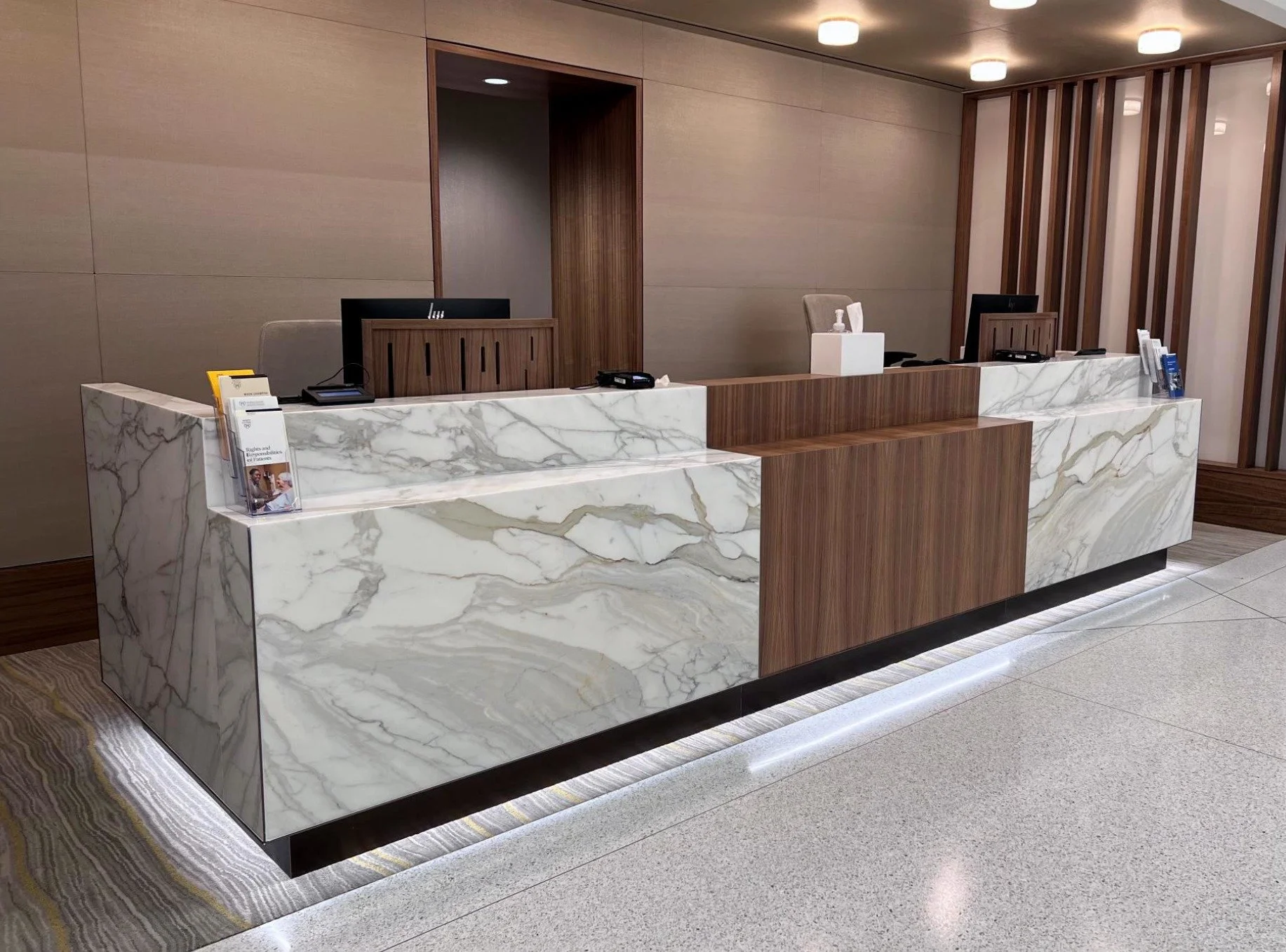 A modern hotel front desk with a marble countertop and wooden accents, including a computer monitor, hand sanitizer, and informational brochures.