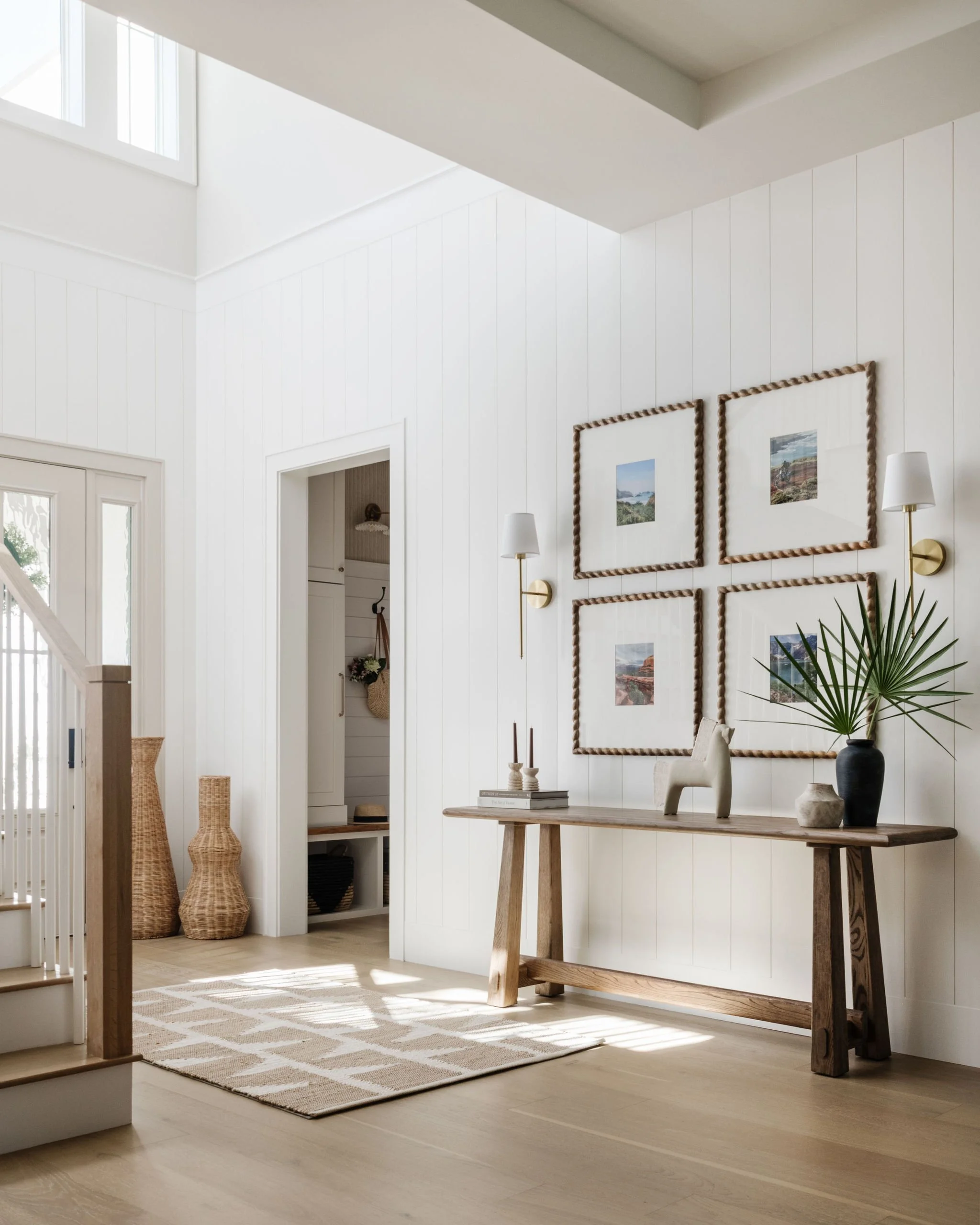 A bright, minimalist living space with white walls and a natural wood floor. Decor includes a rustic wooden table with vases and books, framed landscape photographs with rope borders on the wall, and a white floor lamp. A woven basket and plant are a