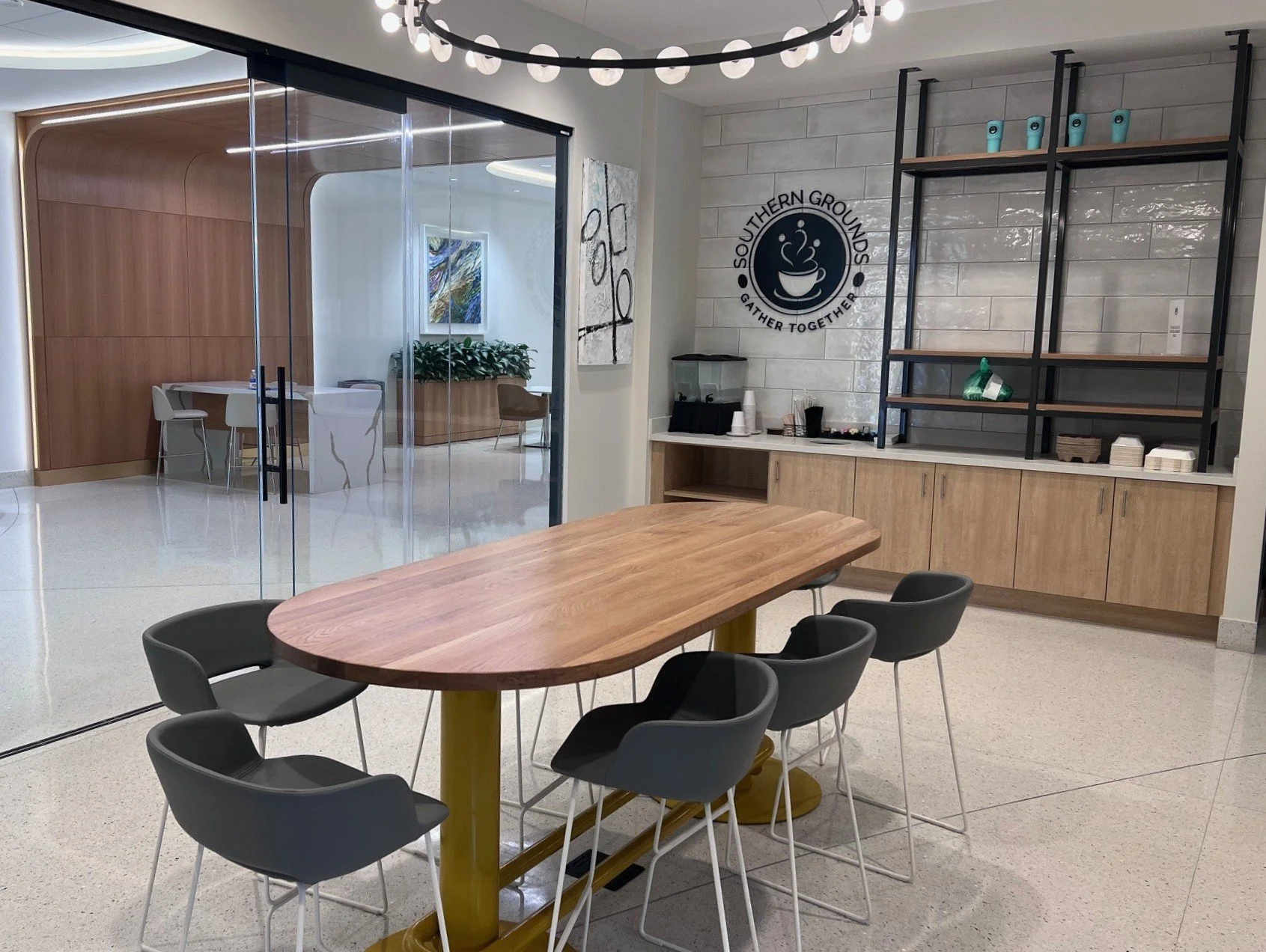 Interior of a modern coffee shop or break area with a wooden table, black chairs, and a counter with shelves, with a logo on the wall that reads 'Southern Grounds Gather Together'.