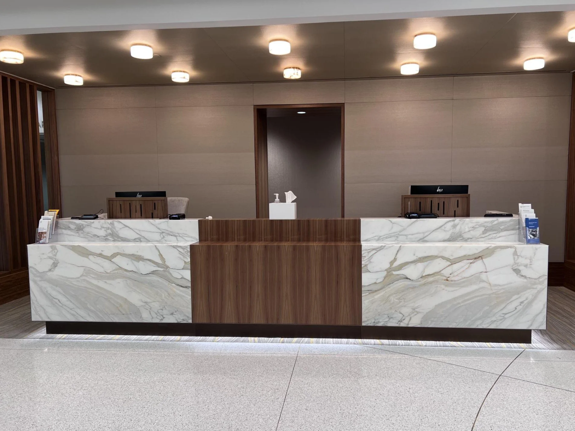 A reception desk made of white marble with gray and gold veining, incorporating dark and light wood accents, situated within a modern interior. The background features a wall with horizontal beige panels and ceiling lights. There are brochures and informational materials on the desk.