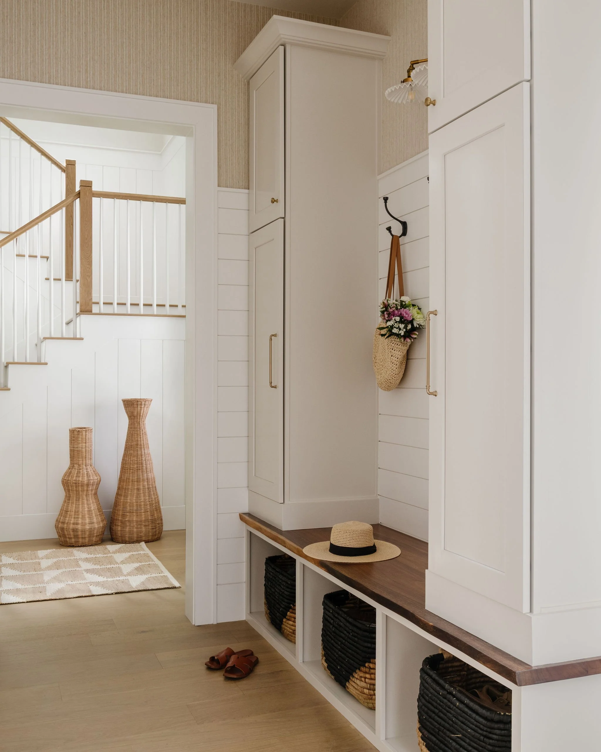 Entryway with white built-in storage, black woven baskets, a straw hat, pink and white flowers in a woven basket hanging on the wall, wicker vases on the floor, and a staircase with wooden handrail in the background.