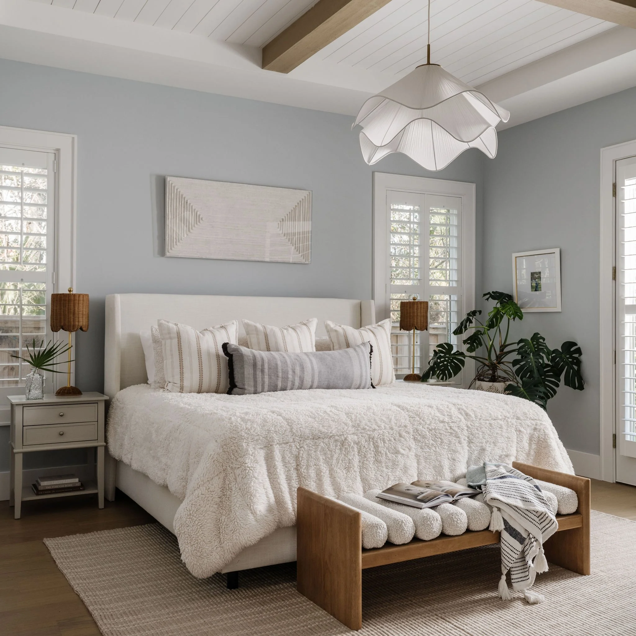 A cozy bedroom with a white fluffy bed, striped pillows, and a wooden bench at the foot of the bed. Two bedside tables with lamps and decorative items. Large windows with white shutters and artwork on the walls. A ceiling light fixture overhead.