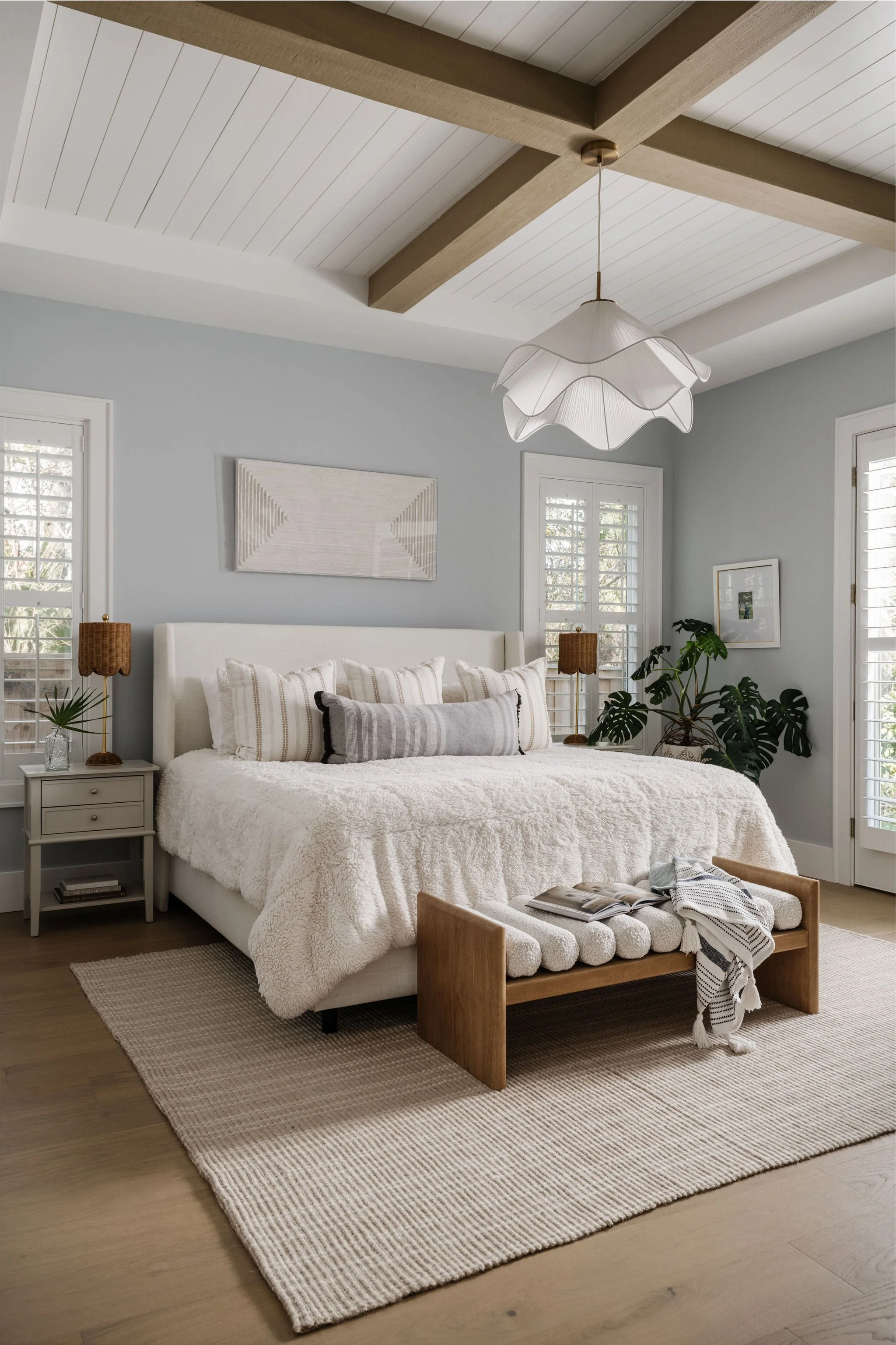 A bedroom with a white bed, beige textured throw, striped pillows, and a wooden bench at the foot. Two side tables with lamps and decor, large windows with white shutters, and a wall art piece. The ceiling features exposed wooden beams and a white pe
