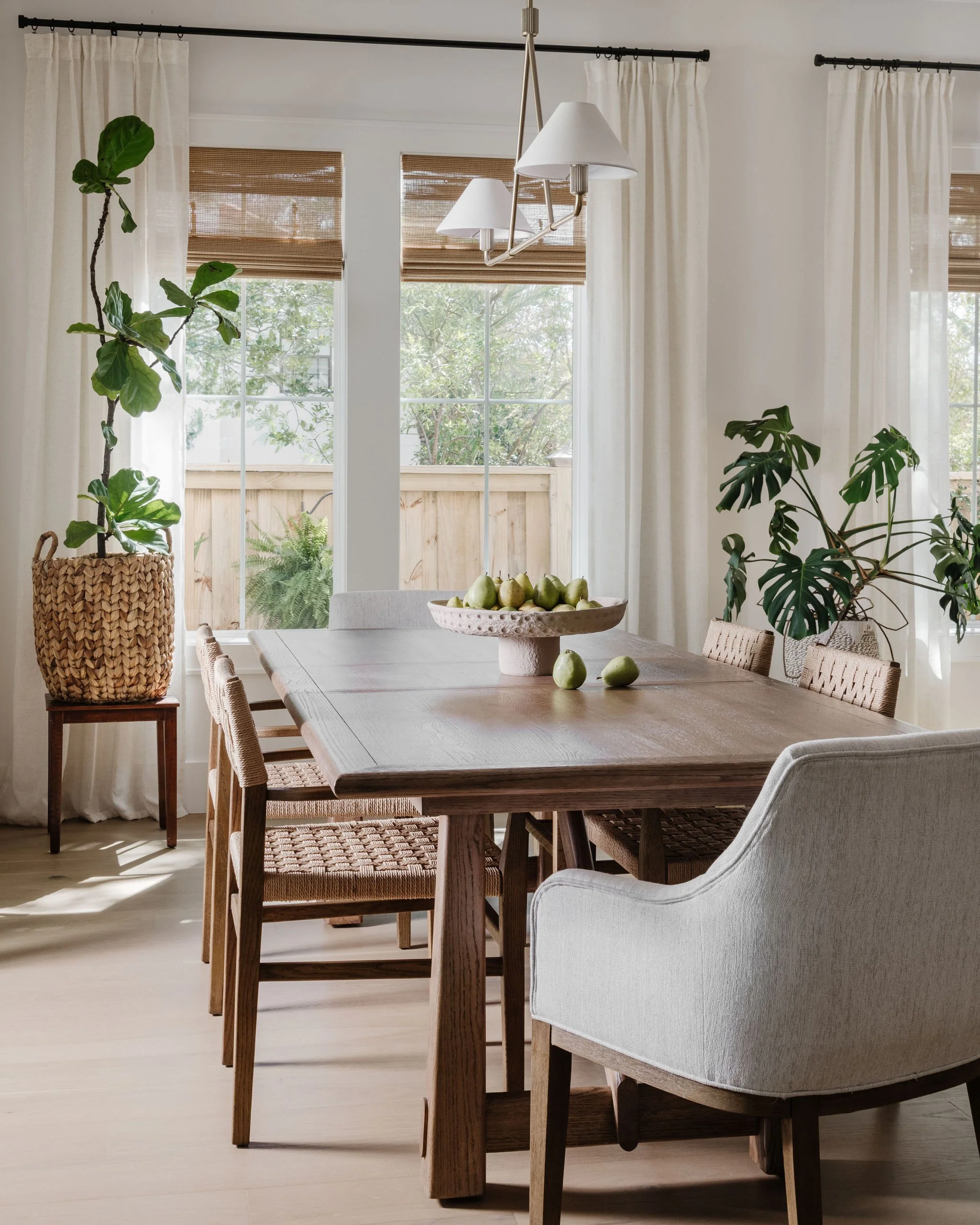Sunlit dining room with a wooden table, white curtains, houseplants, and view of a backyard through sliding glass doors.