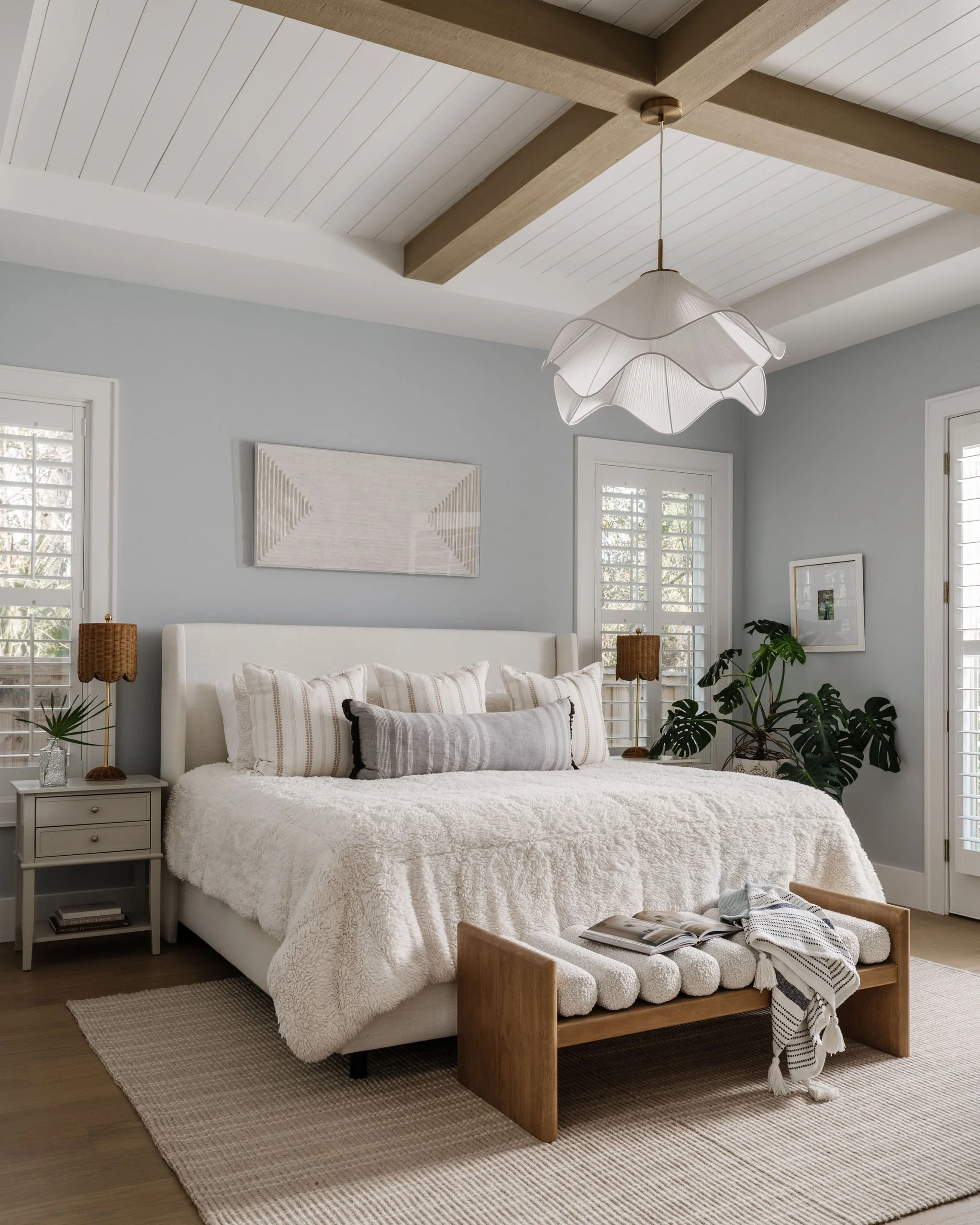 A cozy bedroom with a white bed, beige and white striped pillows, a small side table with magazines, a woven lamp, and green houseplants, with natural light coming through white shutters on windows, and a white ceiling with a wooden beam and a unique