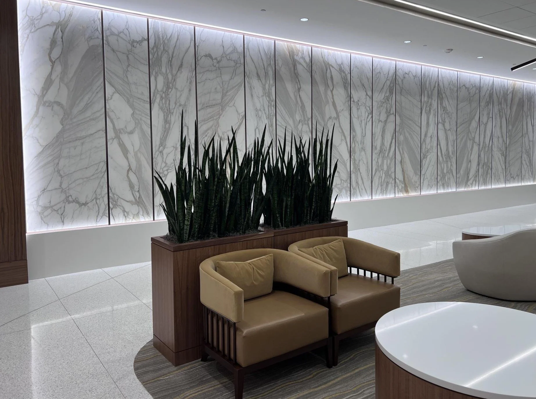 Interior view of a modern lobby with a white marble wall, green indoor plants, beige sofas, and a white circular table.