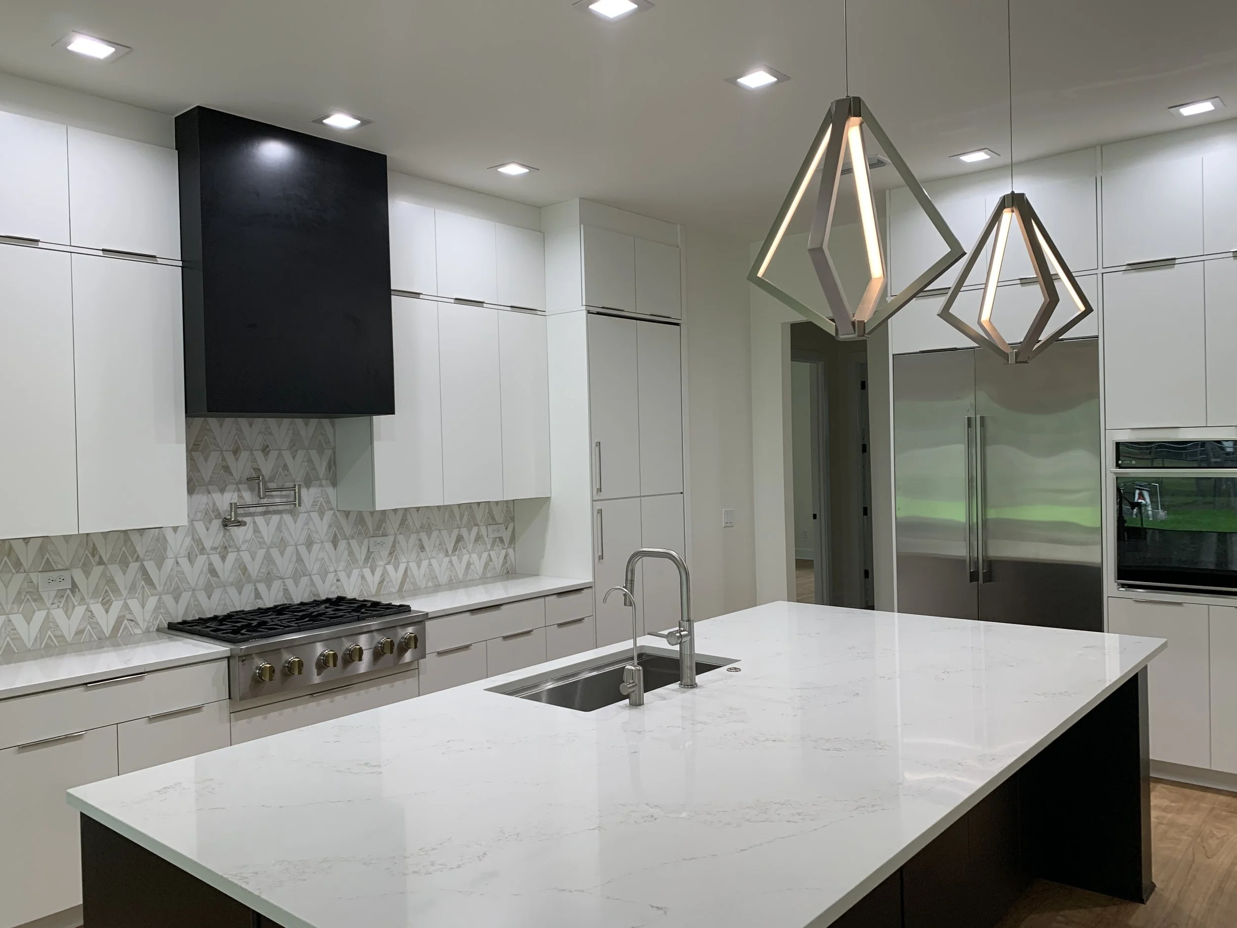 Modern kitchen with white cabinets, a large white island, a stainless steel stove, geometric patterned backsplash, black range hood, and decorative pendant lights hanging from the ceiling.
