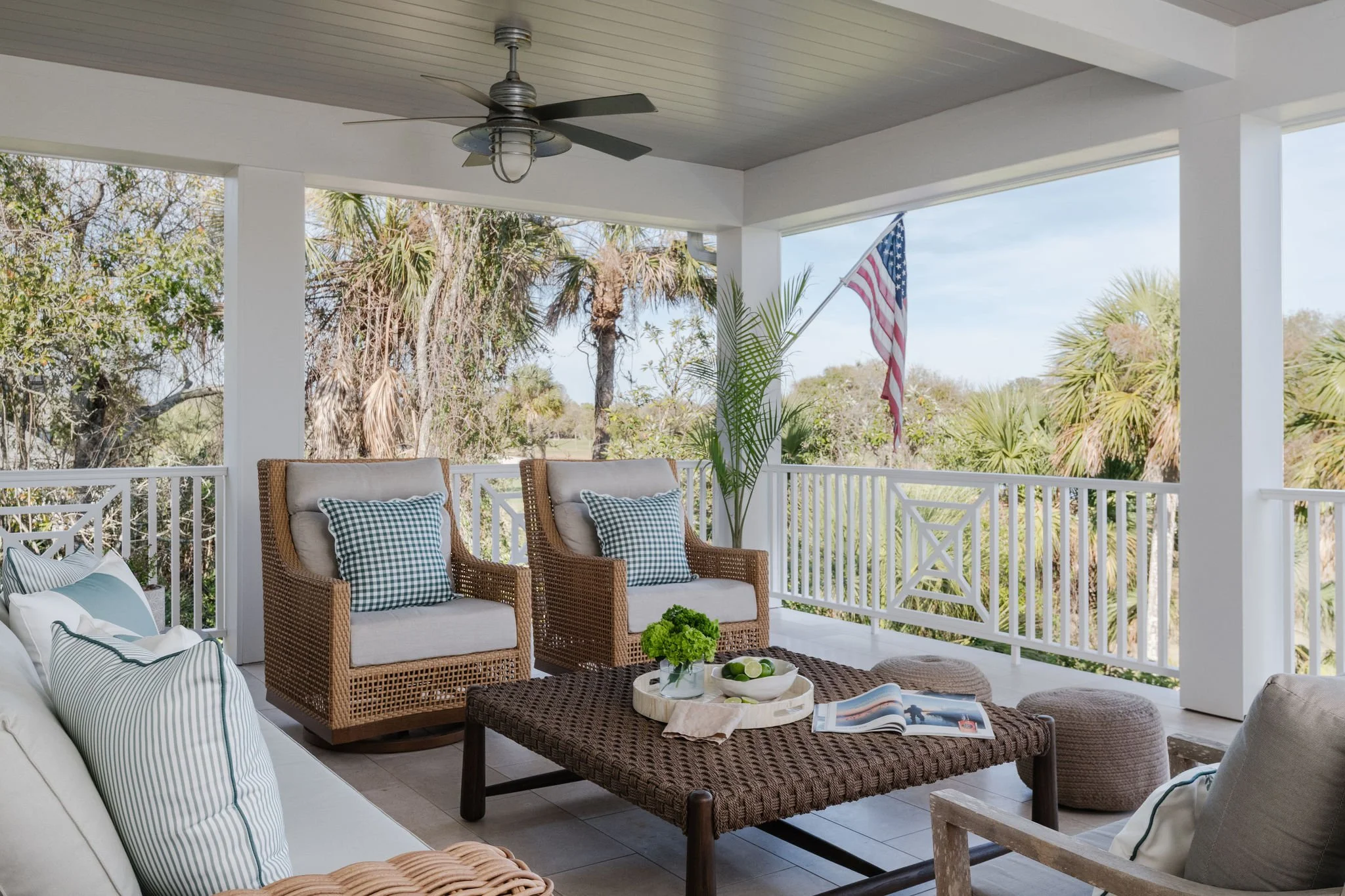 Porch with wicker chairs, cushions, and a coffee table with a magazine, overlooking a backyard with trees and an American flag.