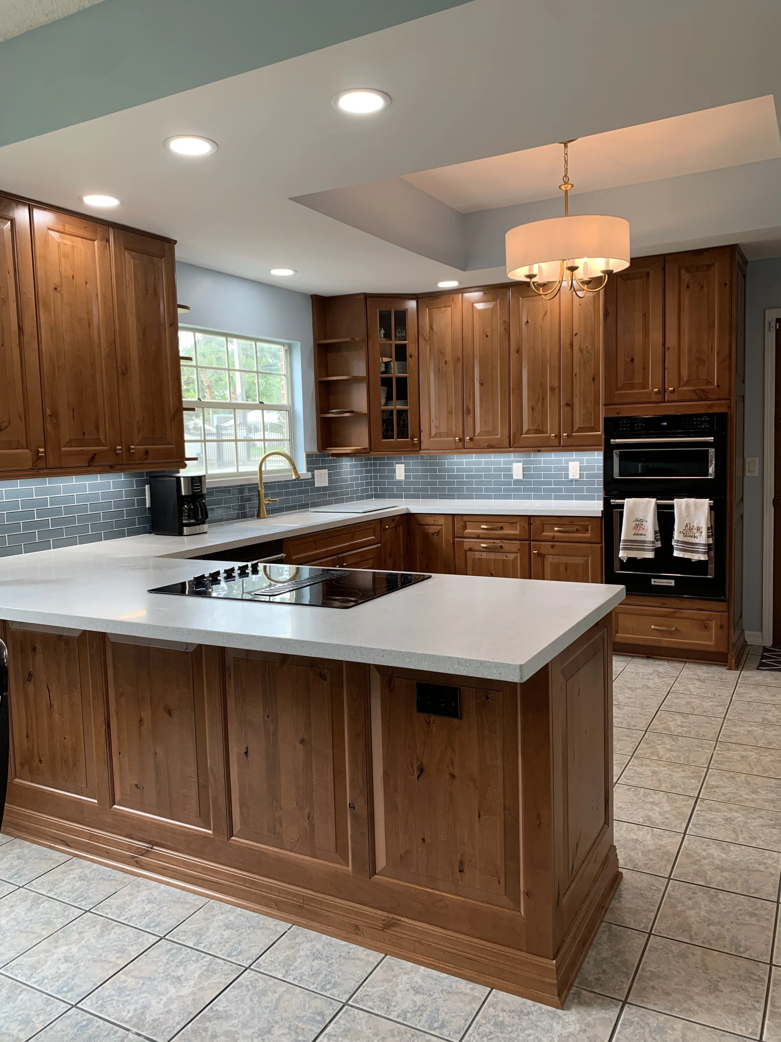 A kitchen with wooden cabinets, a white countertop, a black stove, a window with a view of trees, and a hanging light fixture.