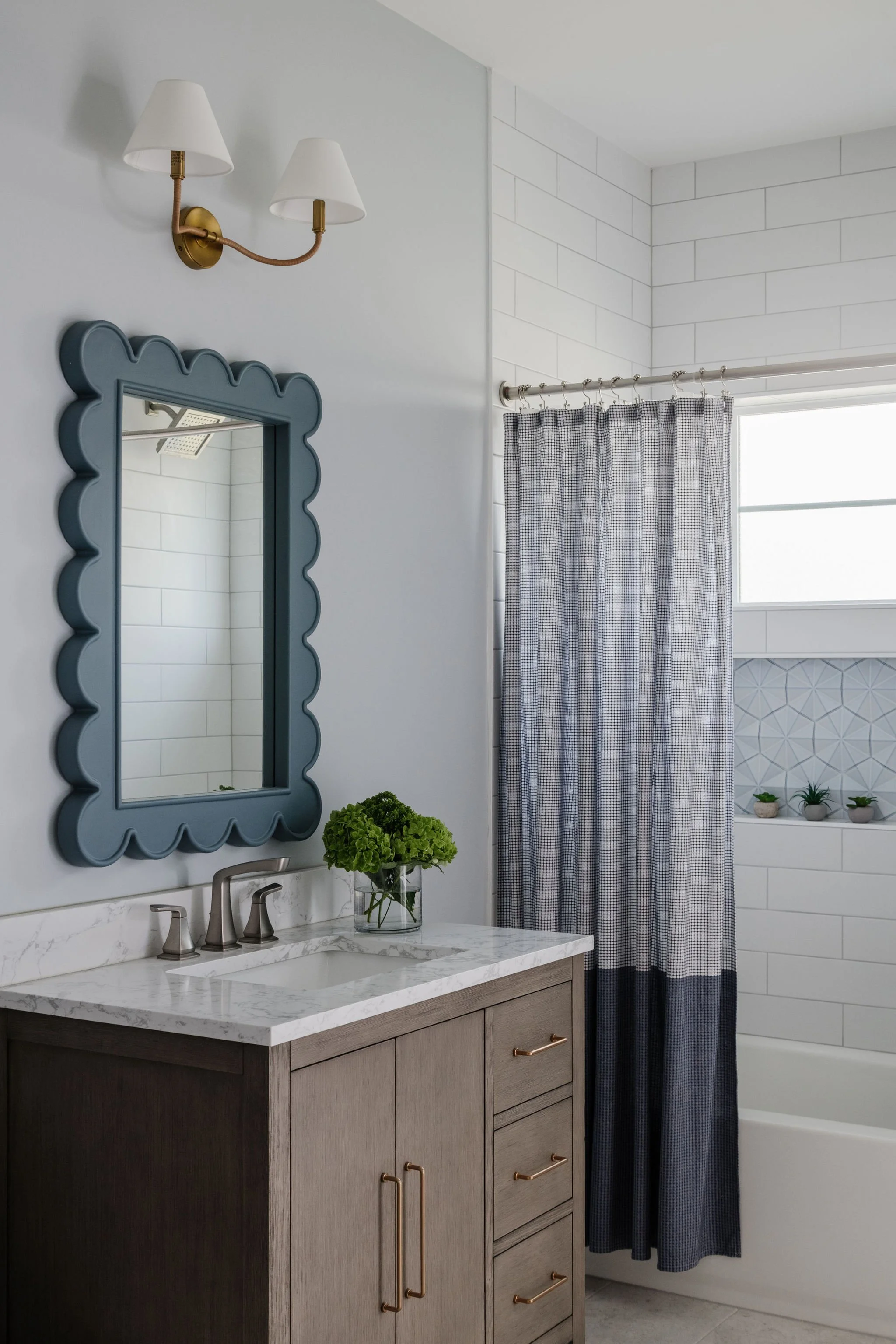 Bathroom with a vanity featuring a marble countertop, a framed mirror with scalloped edges, a small potted plant, and a vase of green hydrangeas. There is a dual-sink faucet, a wall-mounted light fixture with two shades above the mirror, a window wit