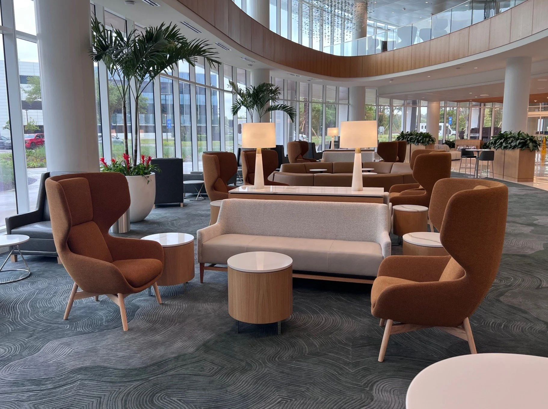 An interior view of a modern airport lounge with numerous comfortable chairs, small tables, large potted plants, and large glass windows allowing natural light, with an upper balcony and seating area.