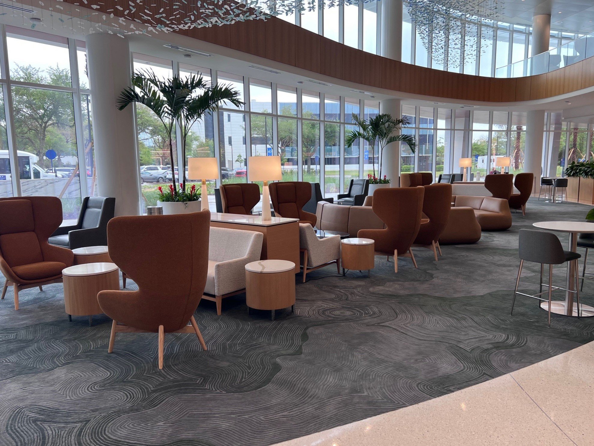 The image shows the interior of a modern, spacious lobby area with large floor-to-ceiling windows, allowing natural light to fill the space. There are several comfortable chairs and small tables arranged throughout the room. Tall indoor plants in whi