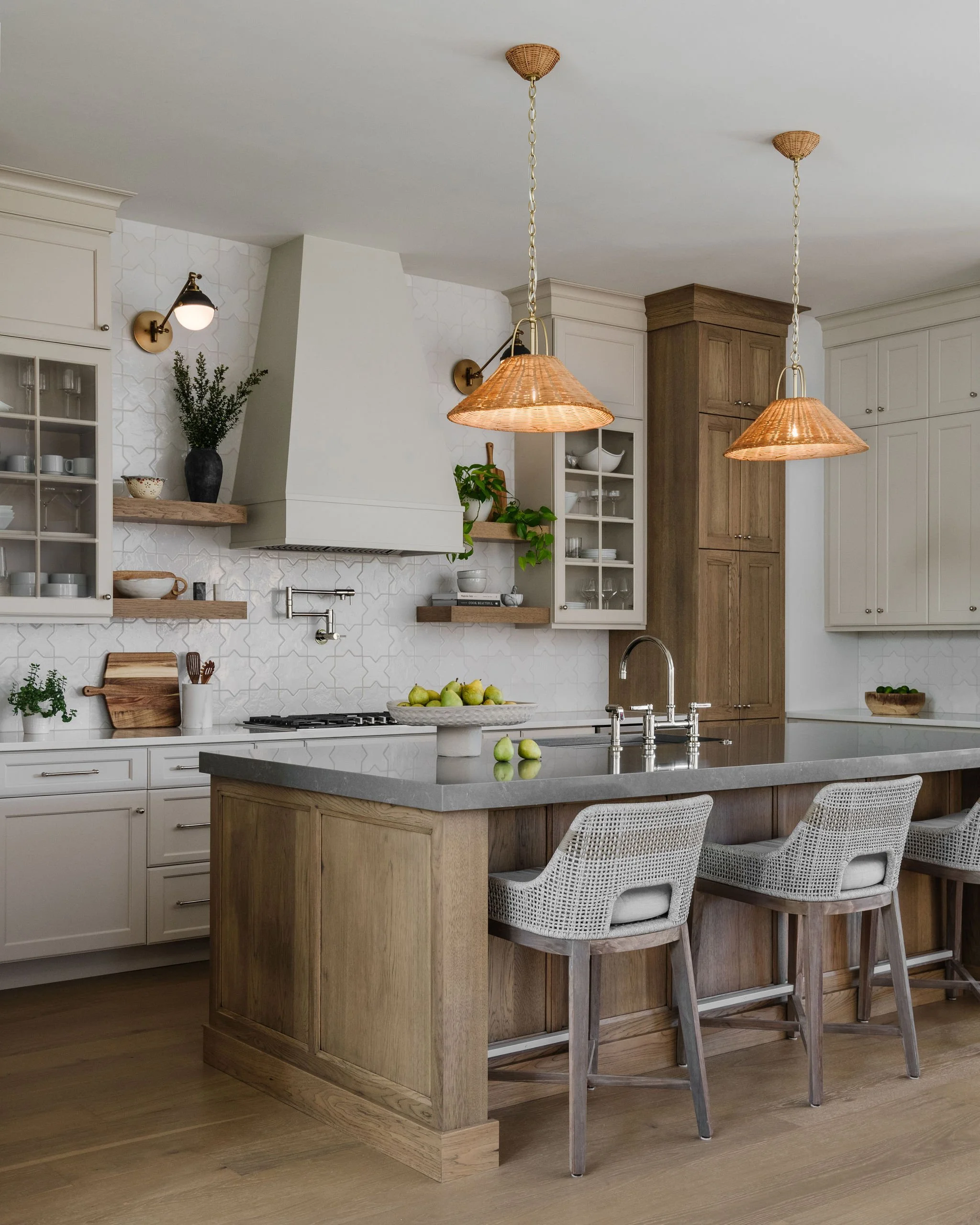 Modern kitchen with white cabinets, wooden accents, and two wicker pendant lights over a kitchen island with a bowl of pears. There are also woven chairs at the island, a wall-mounted lamp, and various kitchenware on open shelves.