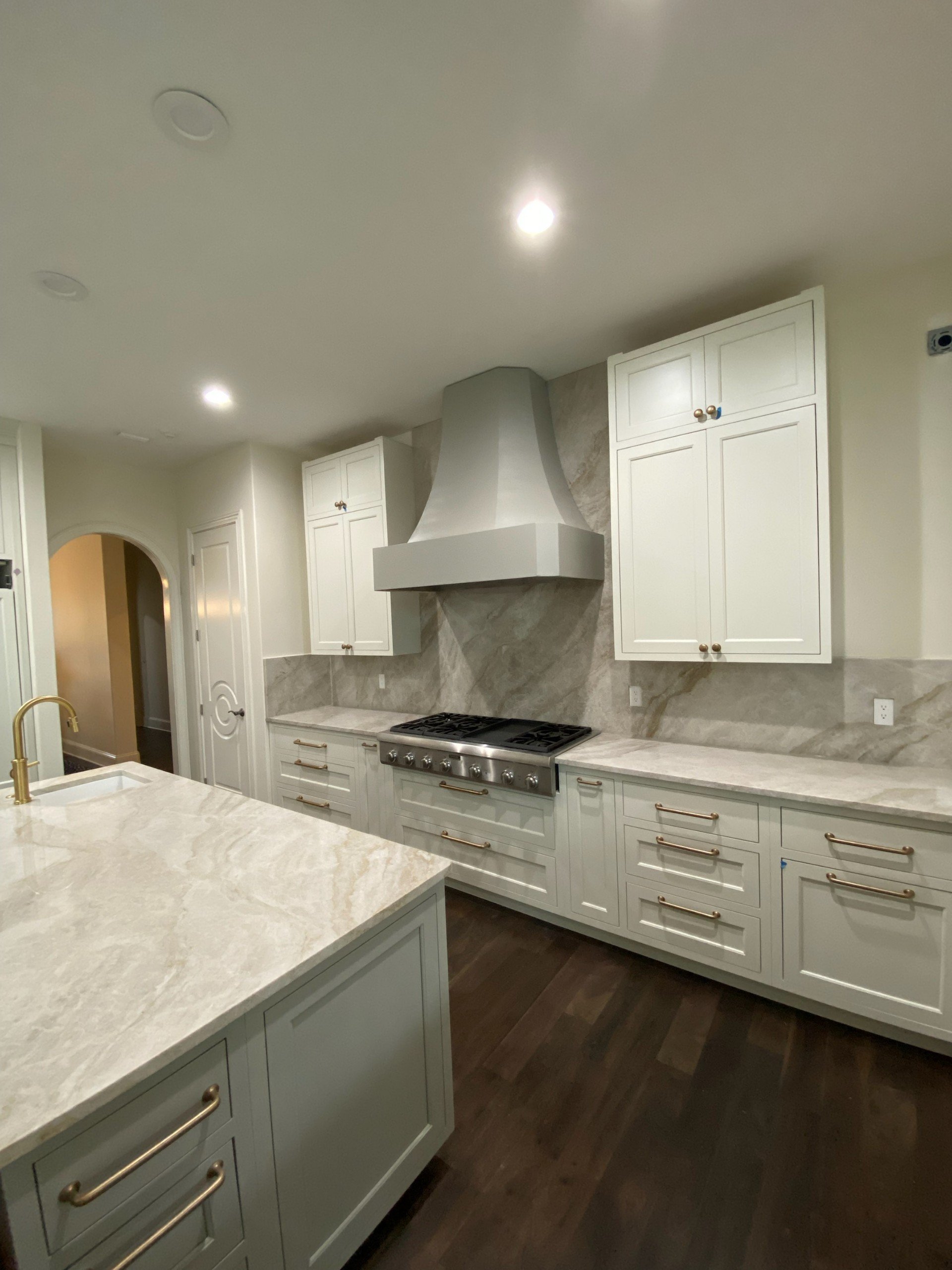 A modern kitchen with white cabinets, a marble countertop, a stainless steel stove, and a marble backsplash. Recessed ceiling lights and a gold faucet are visible.