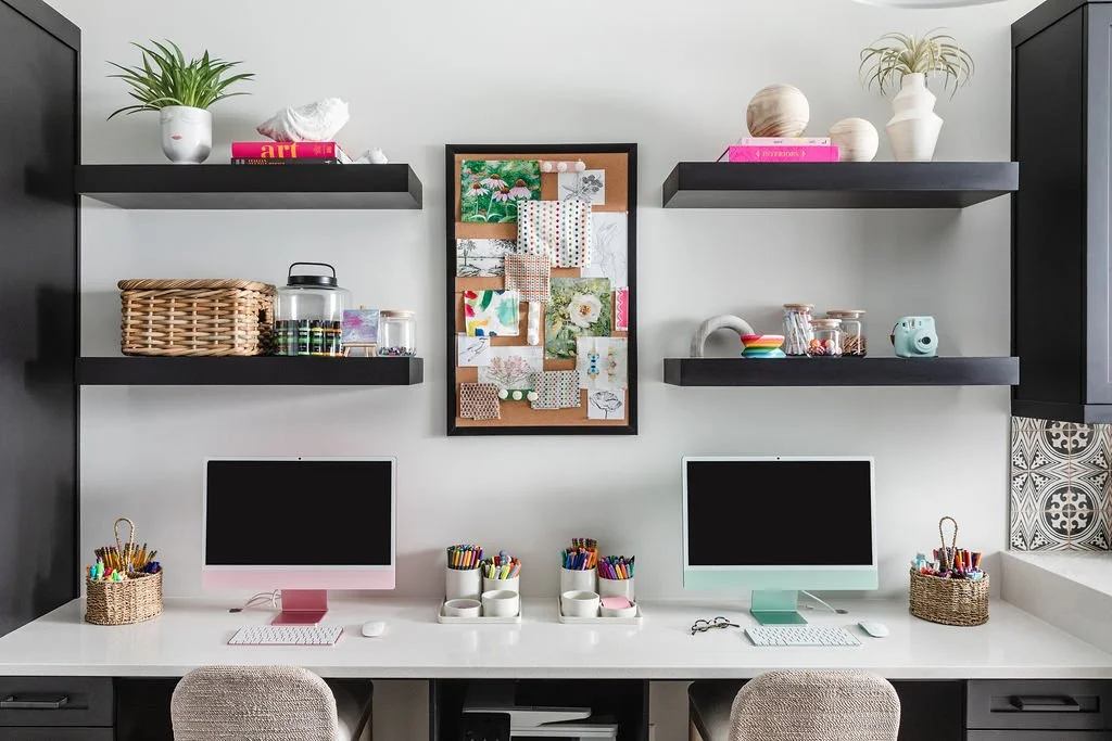 Home office workspace with two desktop computers, colorful pens and supplies, black wall-mounted shelves with decorative items, and a corkboard with photos and fabric swatches.
