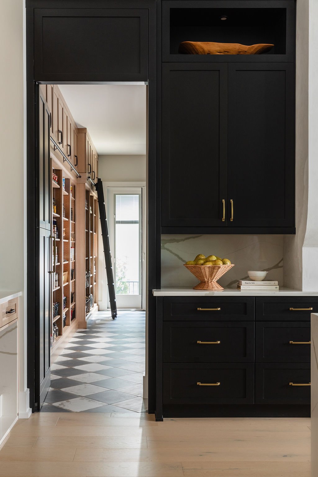 Interior view through a doorway showing a black cabinet with gold handles, a marble backsplash, a woven bowl with lemons, and a white countertop. Beyond, a room with wooden wall shelves, a glass door, and a ladder.