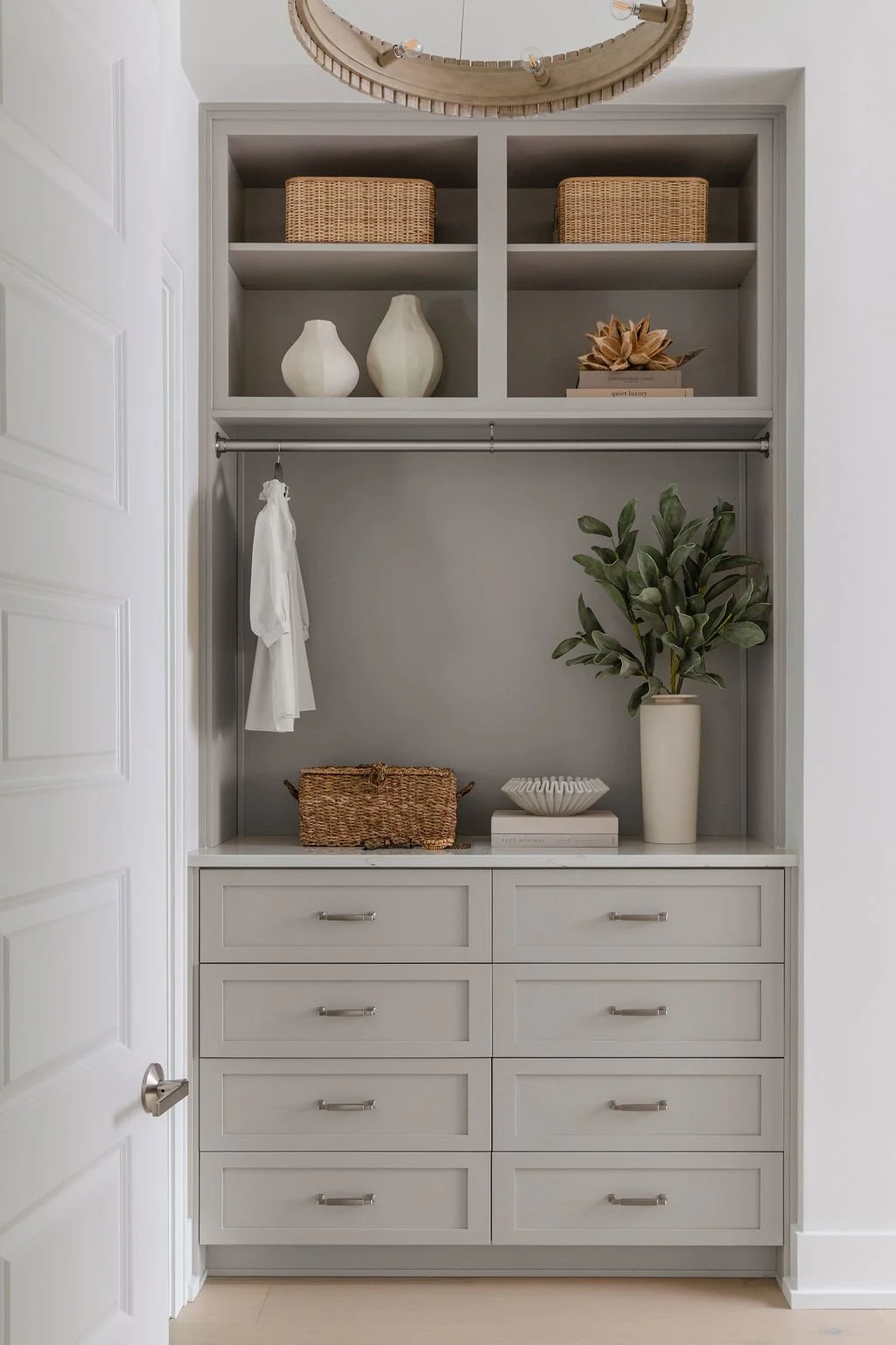 A built-in gray wardrobe with open shelves and six drawers. The shelves contain wicker baskets, white vases, dried flowers, and books. There is a hanging rod with a white piece of clothing and a tall potted plant with green leaves and a cream-colored