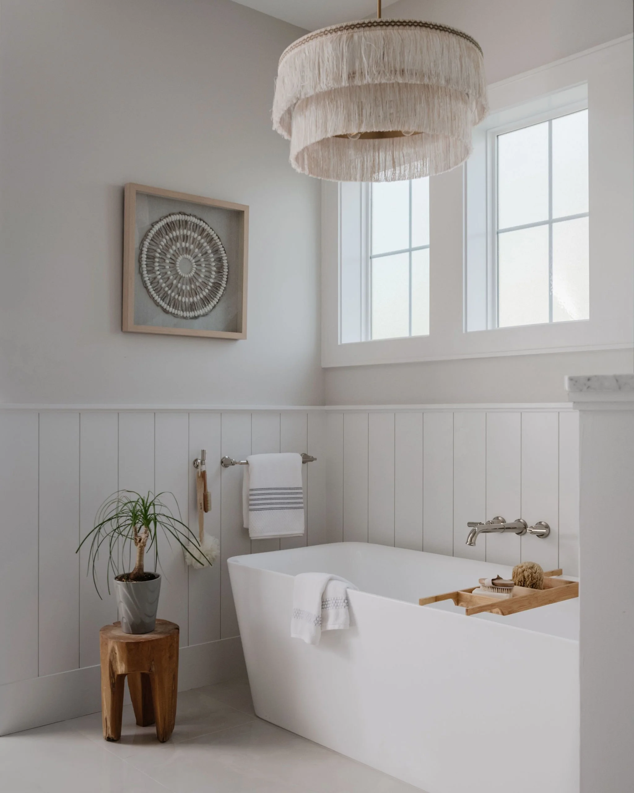 A modern bathroom with white wainscoting, a freestanding bathtub, a wooden stool with a potted plant, and a framed piece of wall art.