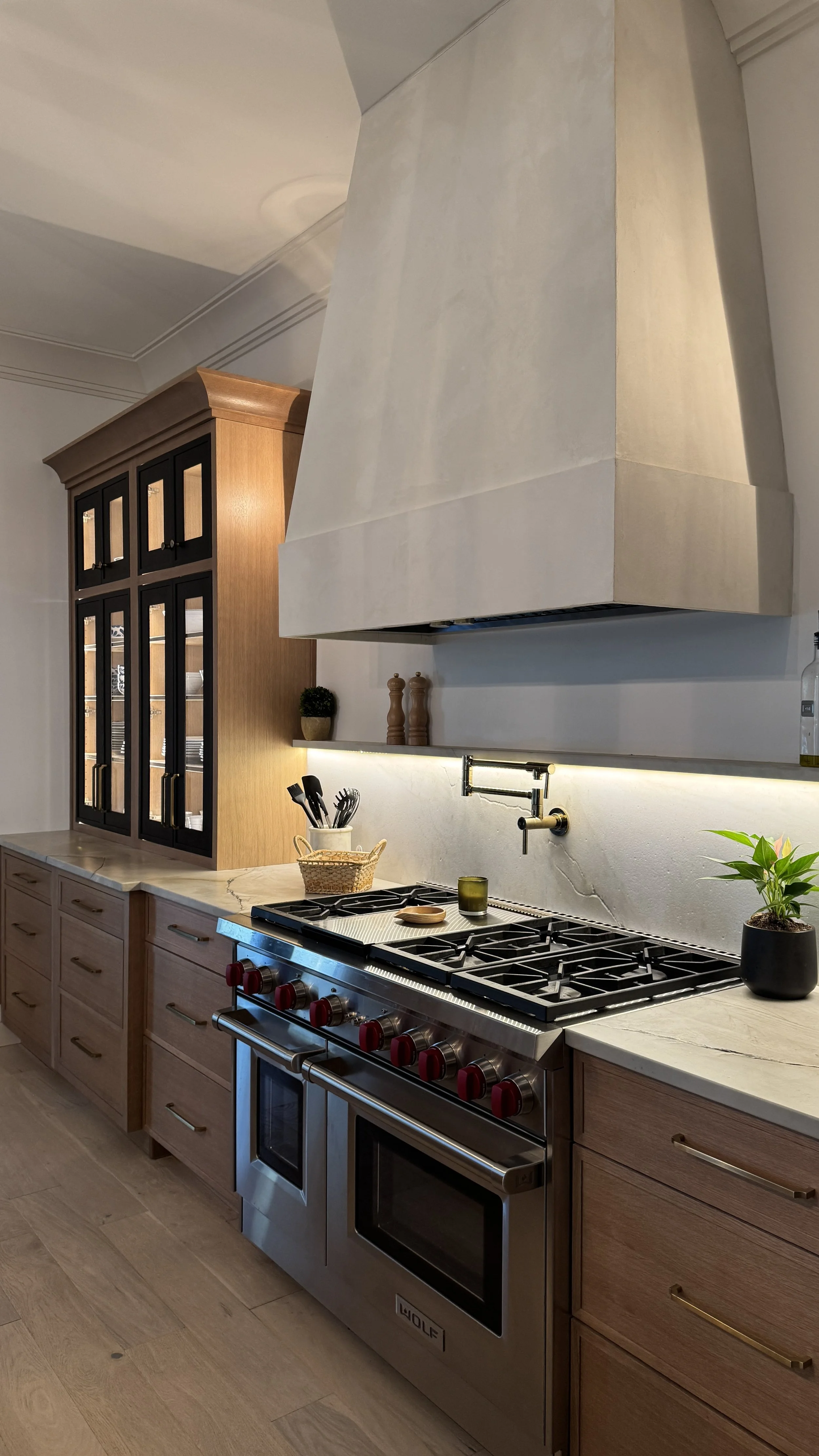 Modern kitchen with wooden cabinets, a stainless steel Wolf oven, a white marble countertop, and a large beige range hood. There are small potted plants, kitchen utensils, and decorative items displayed.