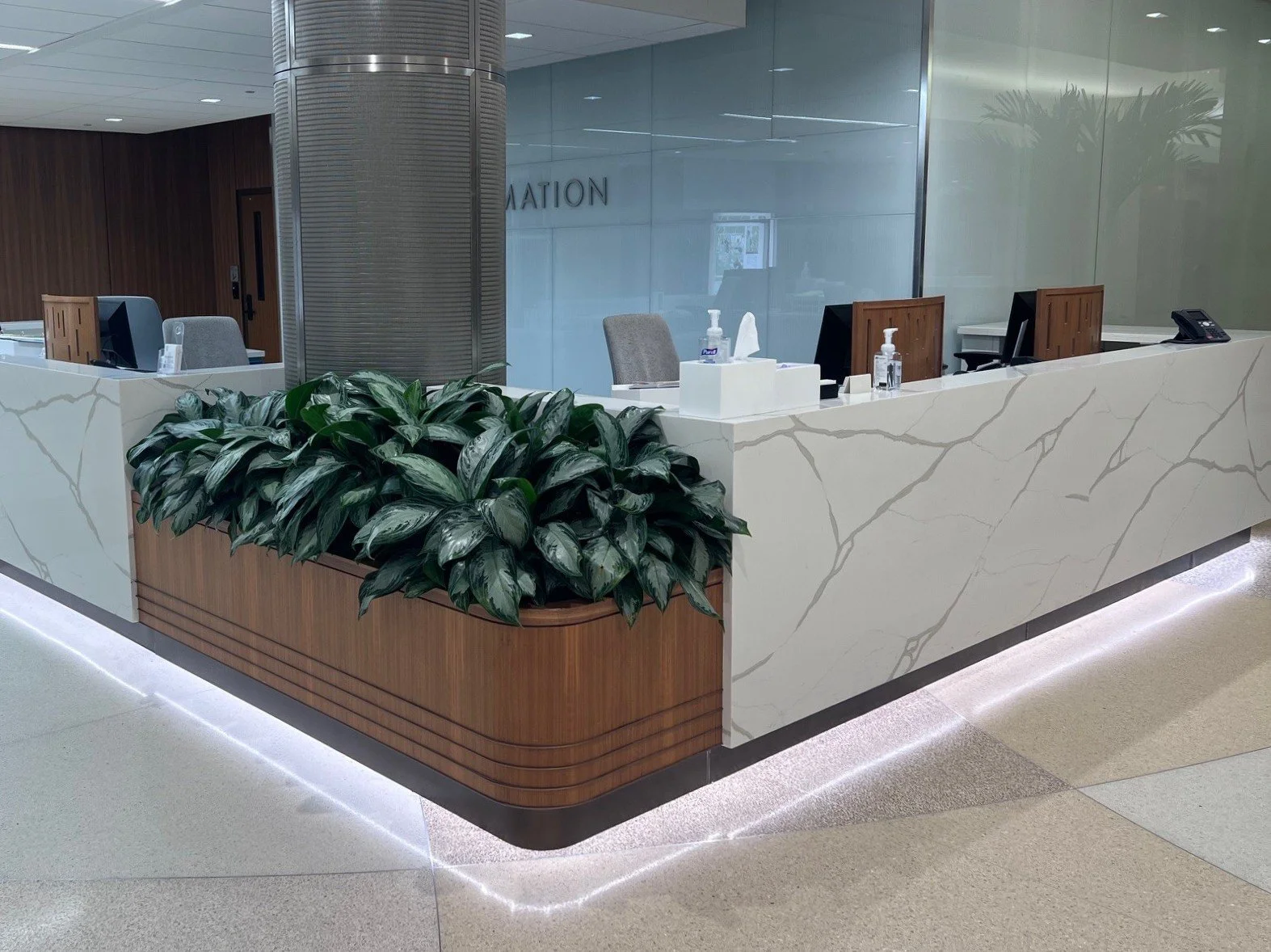 Hospital reception desk with a marble finish, potted green plant, and computers, with a glass wall and elevators in the background.