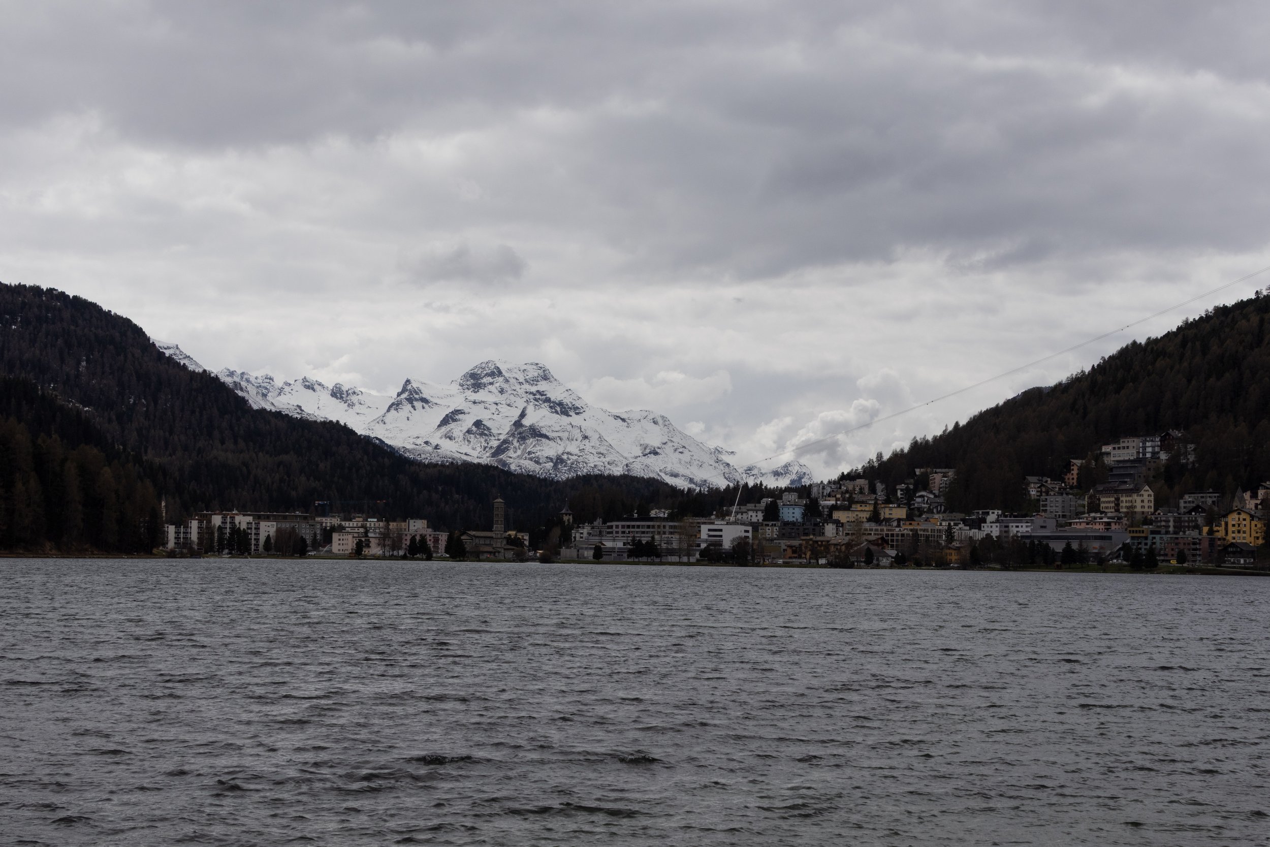 ST. Moritz, Switzerland | Snow-covered mountains rise beyond the lake under an overcast sky. 