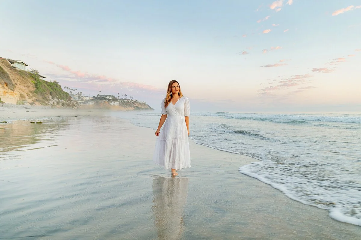 A woman in a white dress walking barefoot along the wet sand on a beach at sunset, with cliffs and houses in the background.