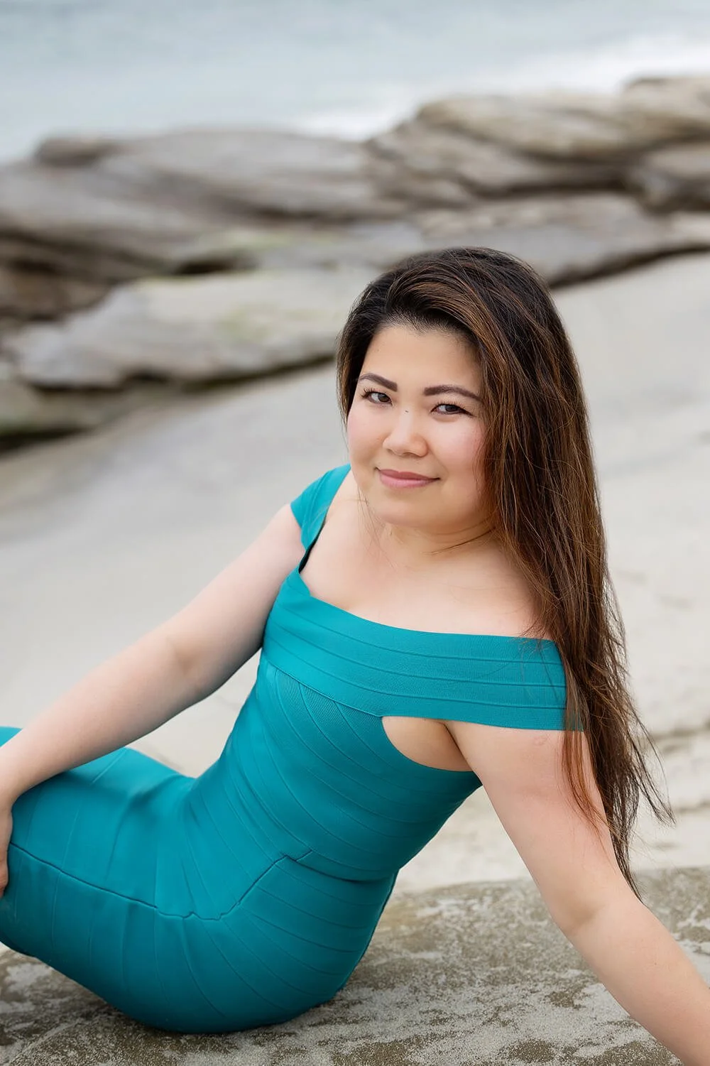 A woman in a teal dress sitting on the sand at the beach, with rocks and water in the background.