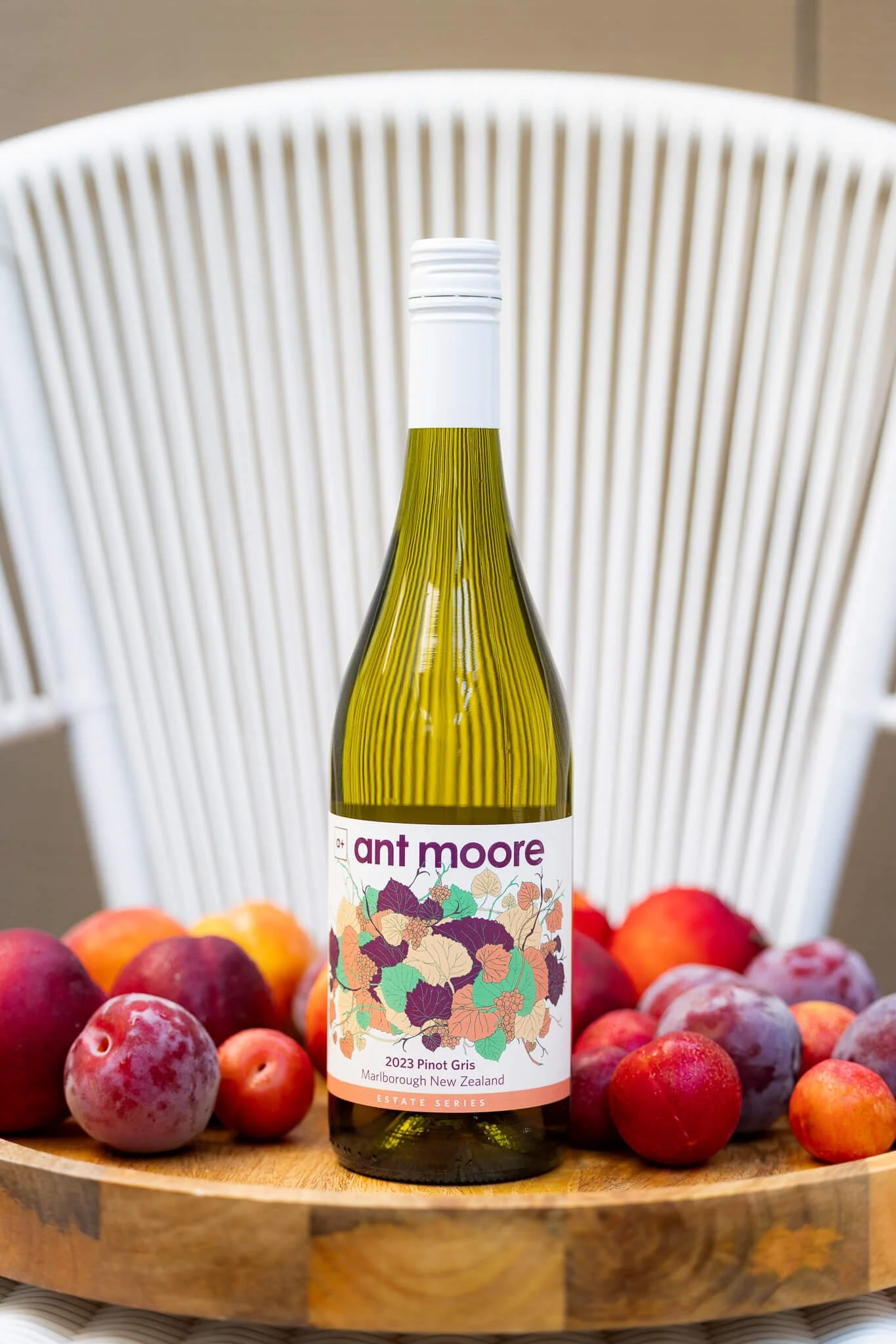A bottle of Ant Moore 2023 Pinot Gris wine from Marlborough New Zealand placed on a round wooden surface surrounded by red and purple grapes and some peaches or nectarines. In the background, there is a white modern chair.